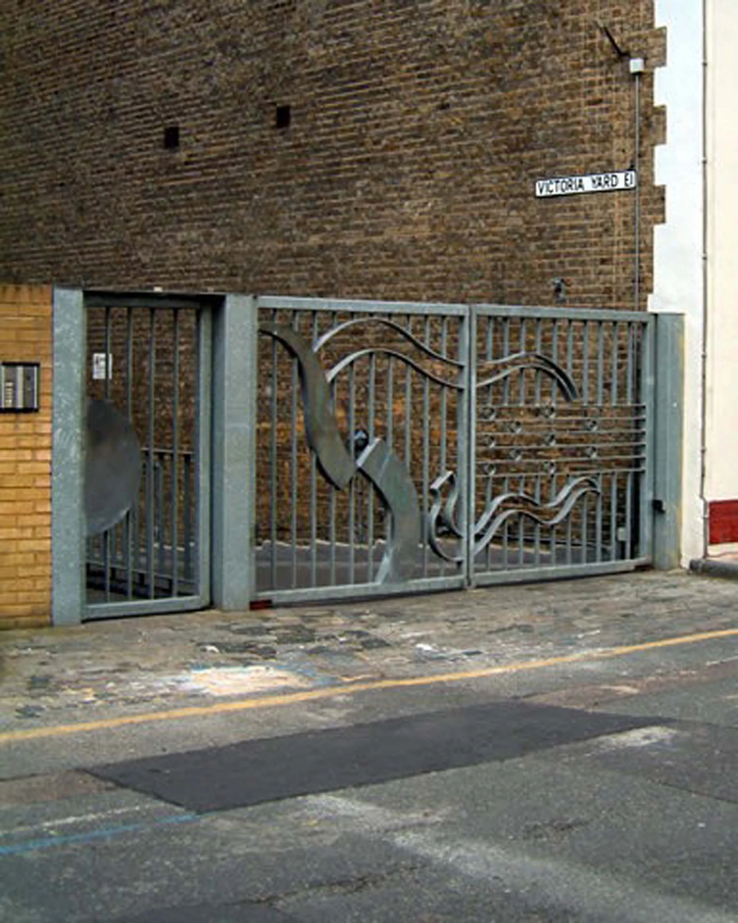 A brick wall with a white street sign that reads "Victoria Yard E" attached to the corner. In front of the wall, there is a metal gate with decorative elements, and the sidewalk appears worn with visible patches and a double yellow line on the street