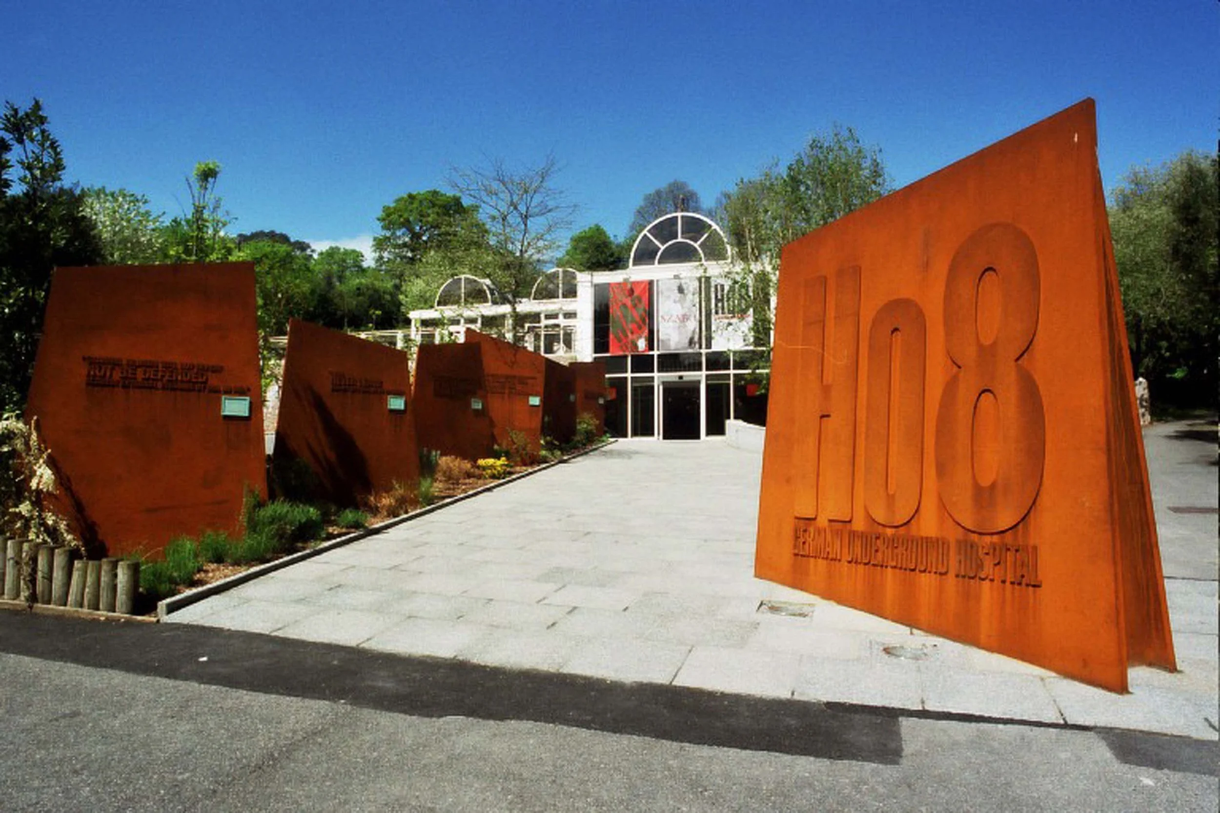 Entrance to the Mexican Underground Museum with large rust-colored metal signs and glass buildings, surrounded by trees and greenery under a blue sky.