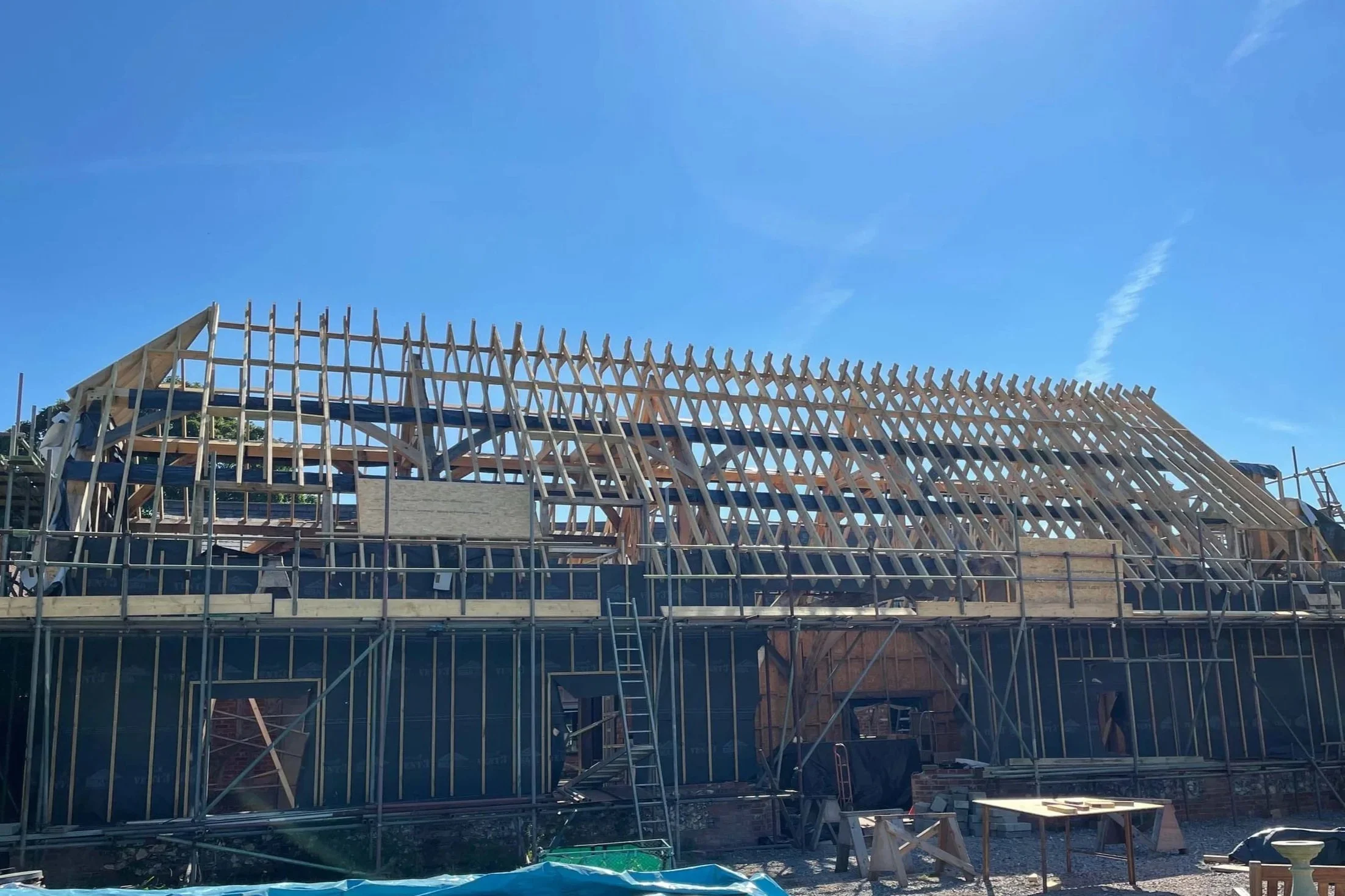 The wooden frame and roof trusses of a house under construction, with scaffolding surrounding the building and blue sky overhead.
