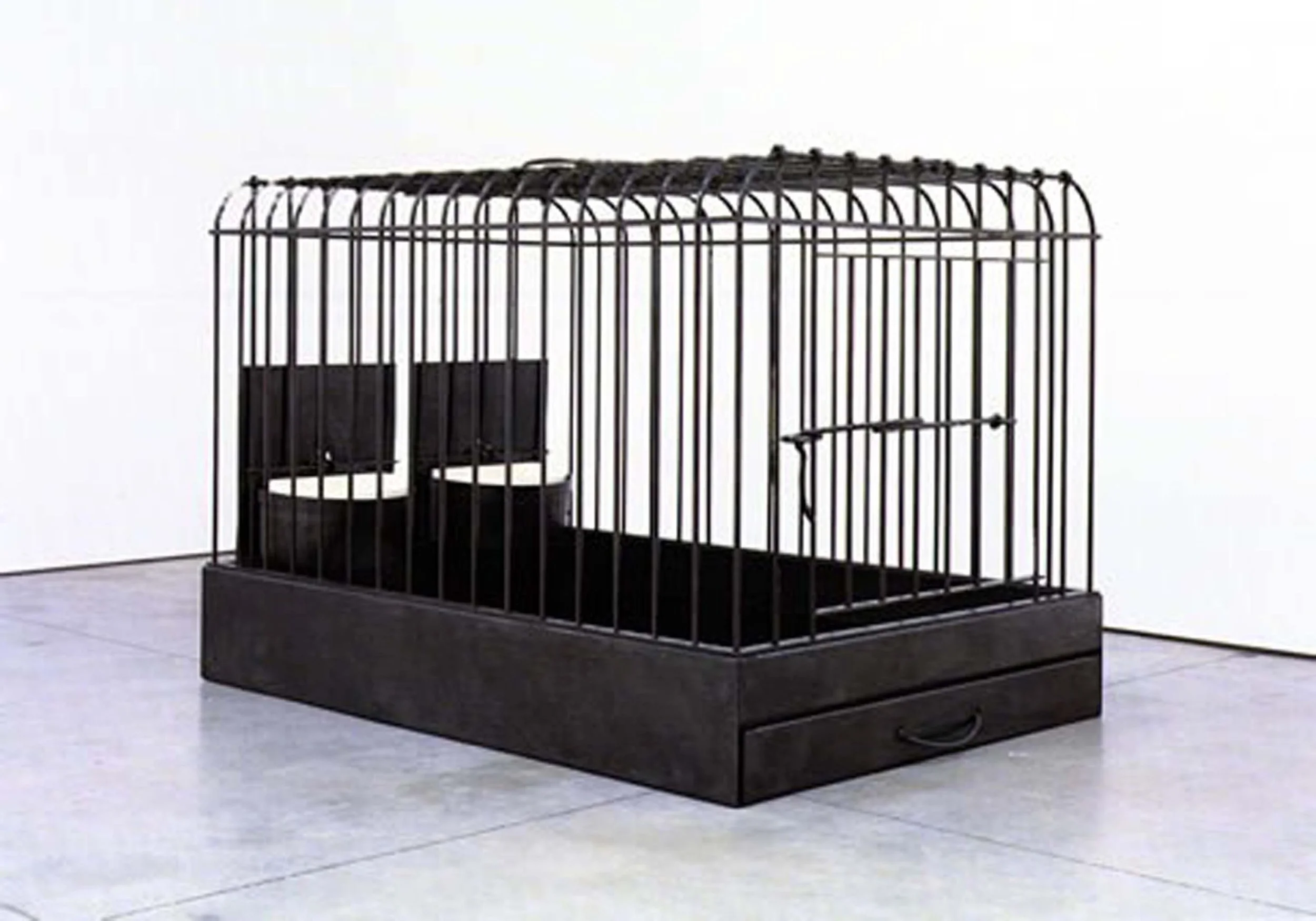 A black decorative birdcage on a black platform inside a gallery or museum space with plain white walls.