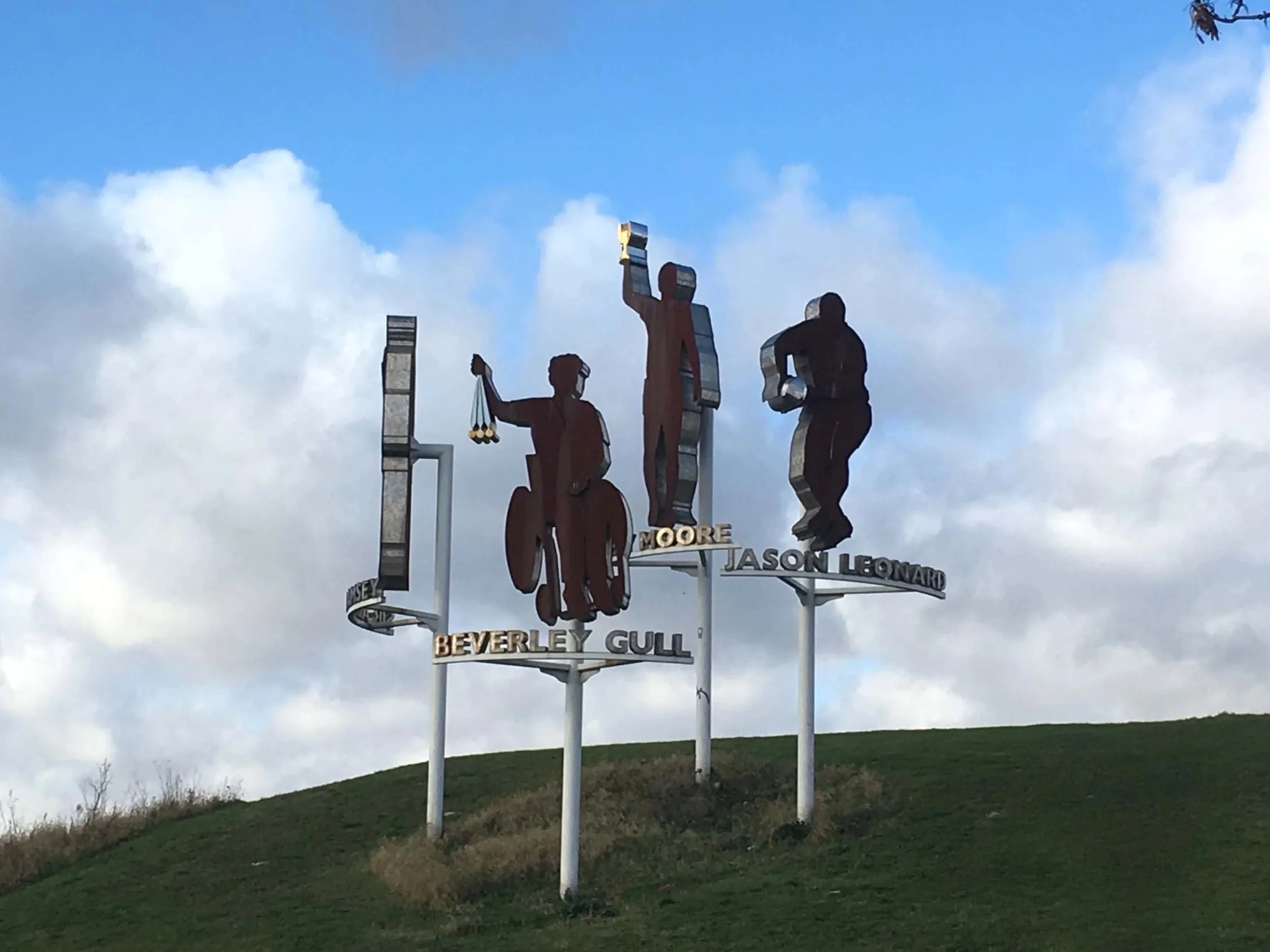 Statue of four human figures with names 'Moore,' 'Jason Leonard,' 'Beverley Gull,' and another name partially visible, set against a cloudy sky and a grassy hill.