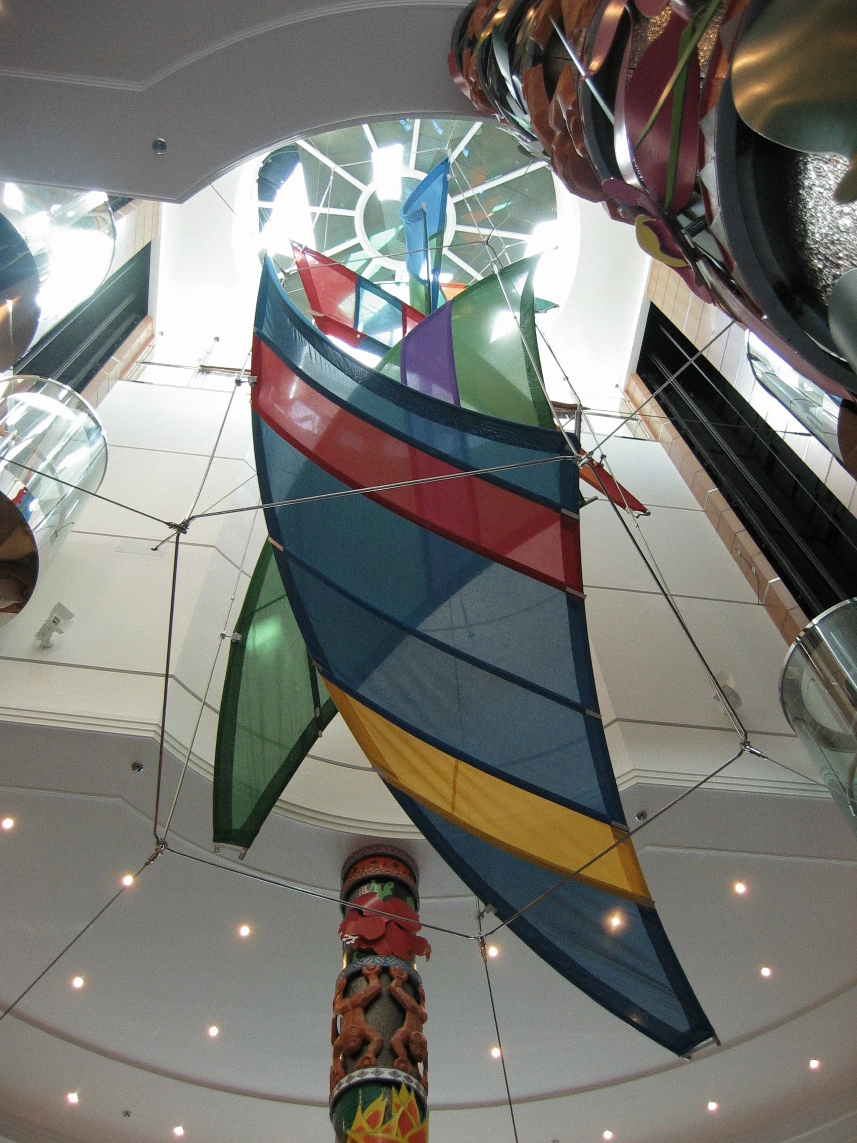 A colorful indoor slide with sections in green, yellow, red, and blue, spiraling around a decorative column with native American style carvings, in a spacious area with a circular ceiling and small ceiling lights.