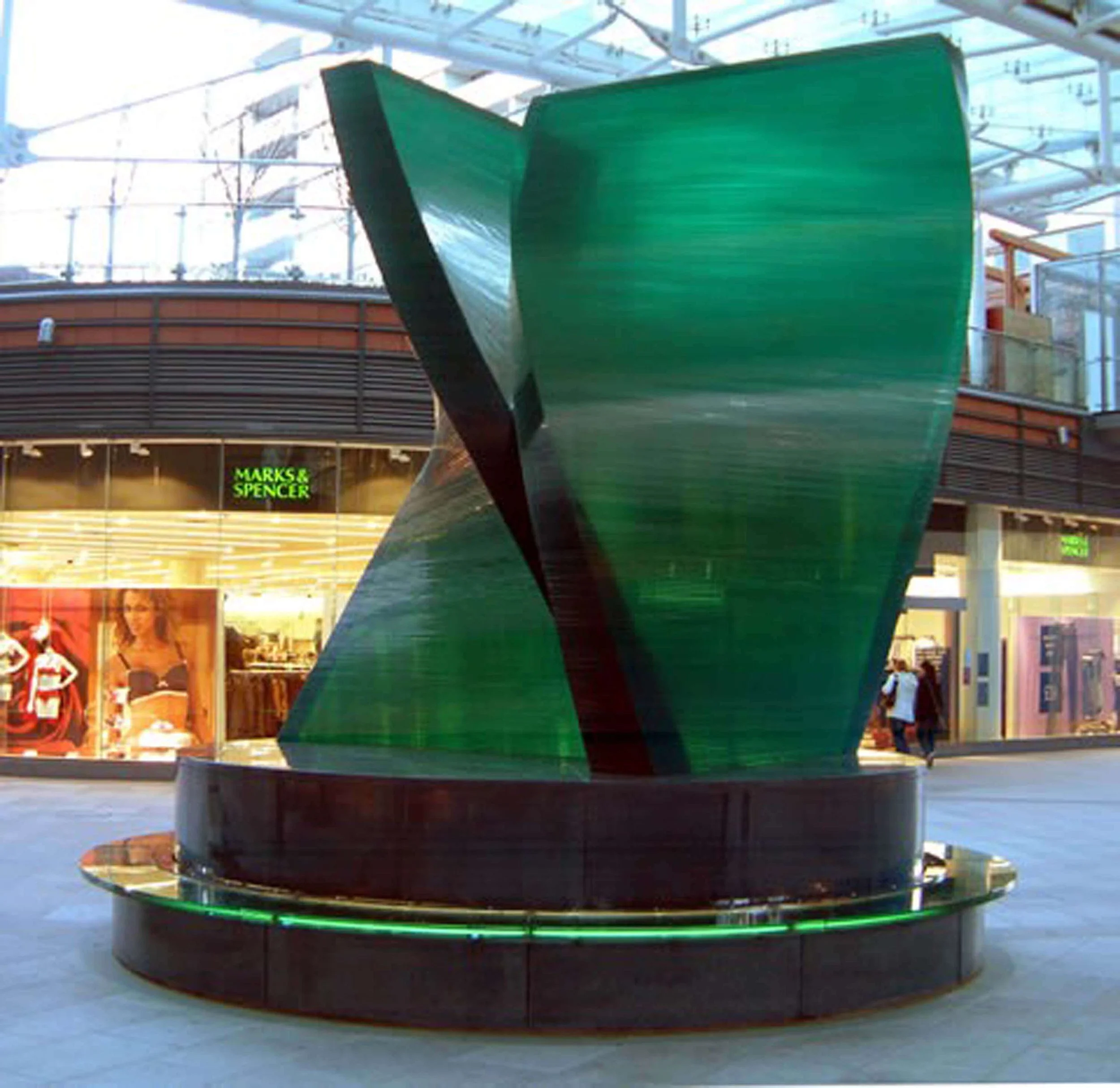 A large, green, ship-shaped sculpture or installation inside a shopping mall. The ship appears to be part of a display or attraction, with a curved, modern design. In the background, there are store signs and shoppers.