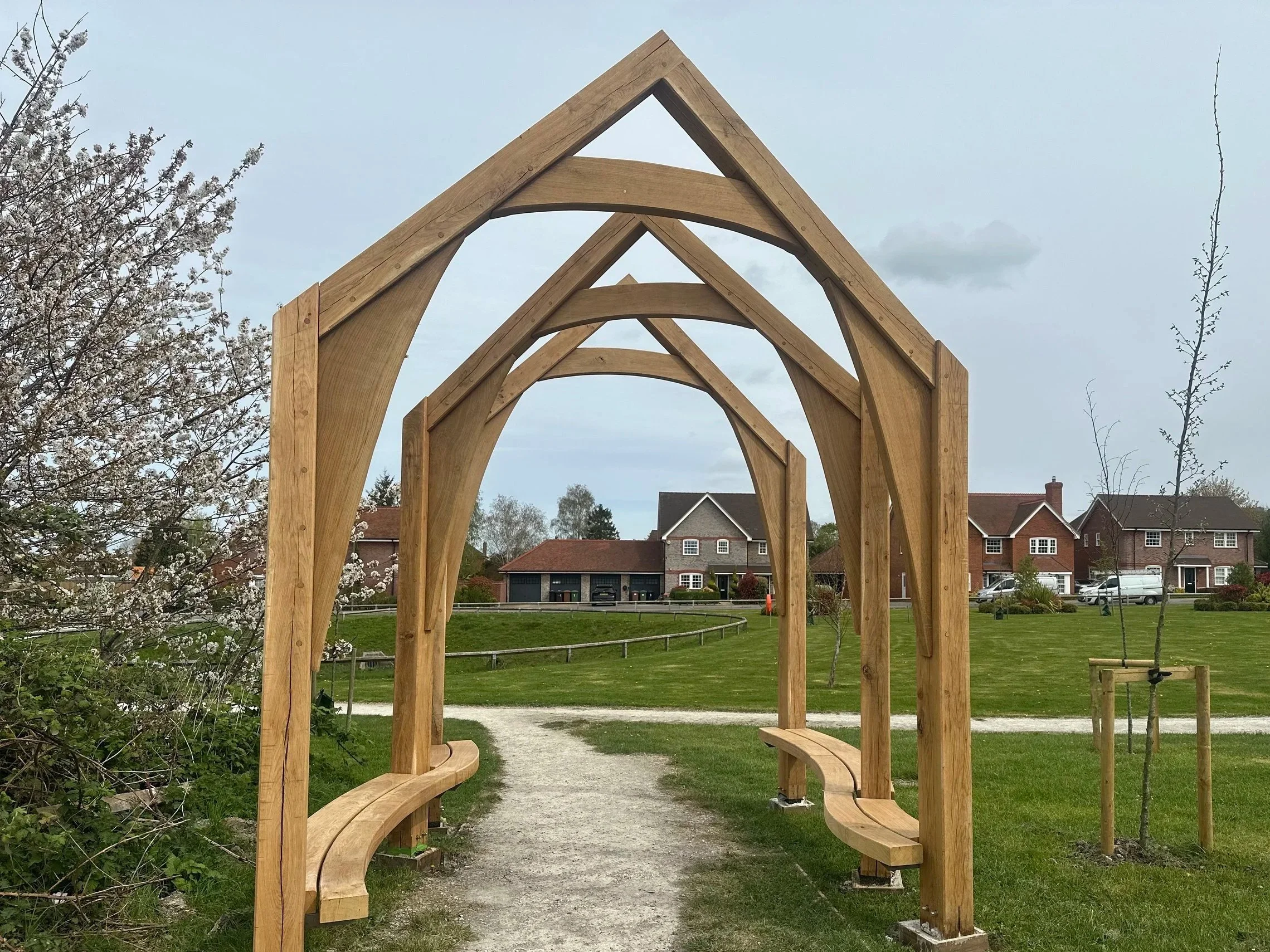 View of a wooden archway with benches on each side, situated in a park with a gravel path, green grass, blooming trees, and houses in the background under a cloudy sky.