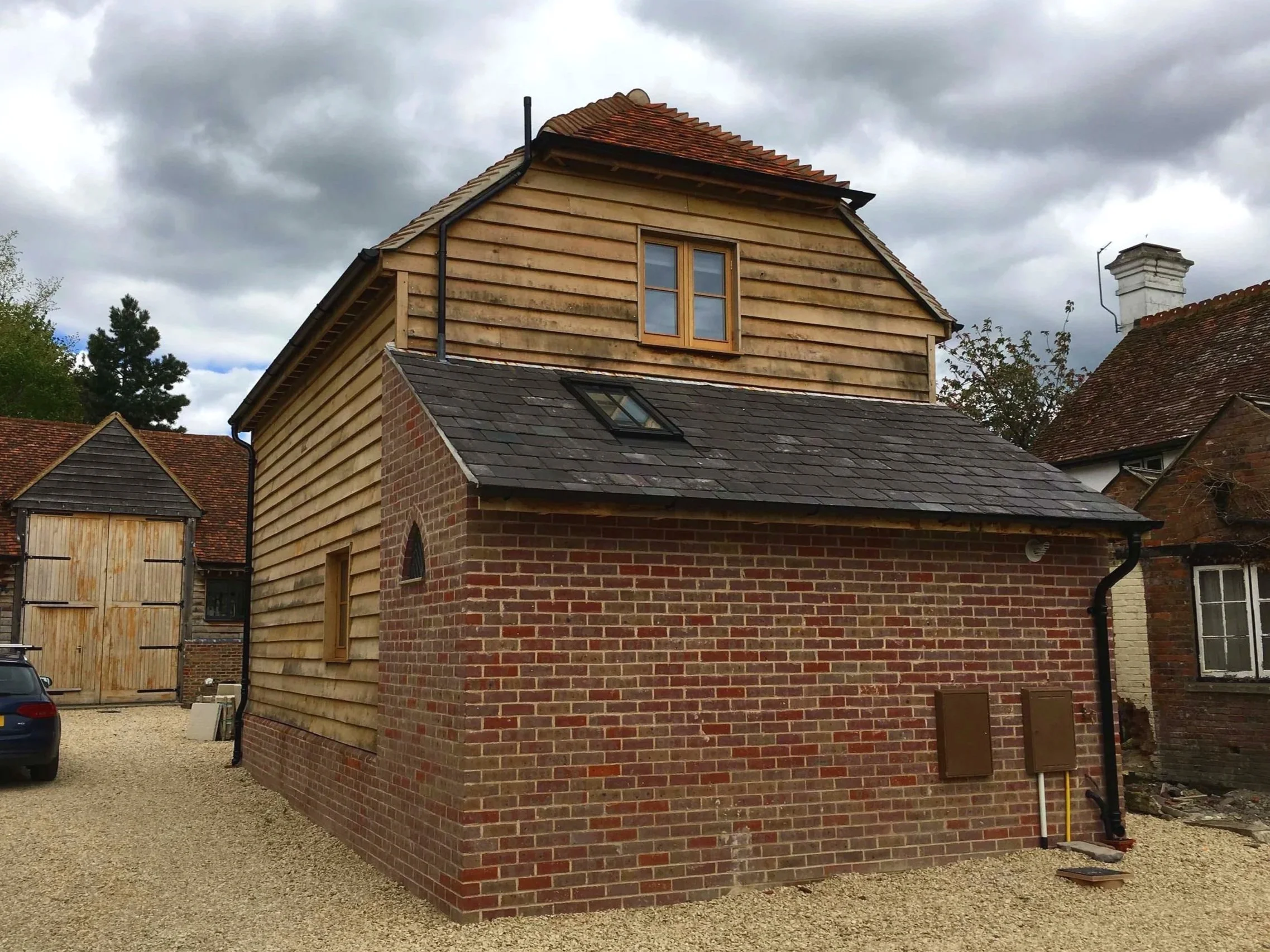 A house with brick and wood siding, a sloped roof with tiles, and a window. There is a gravel driveway and a garage door on the left side. The sky is cloudy.