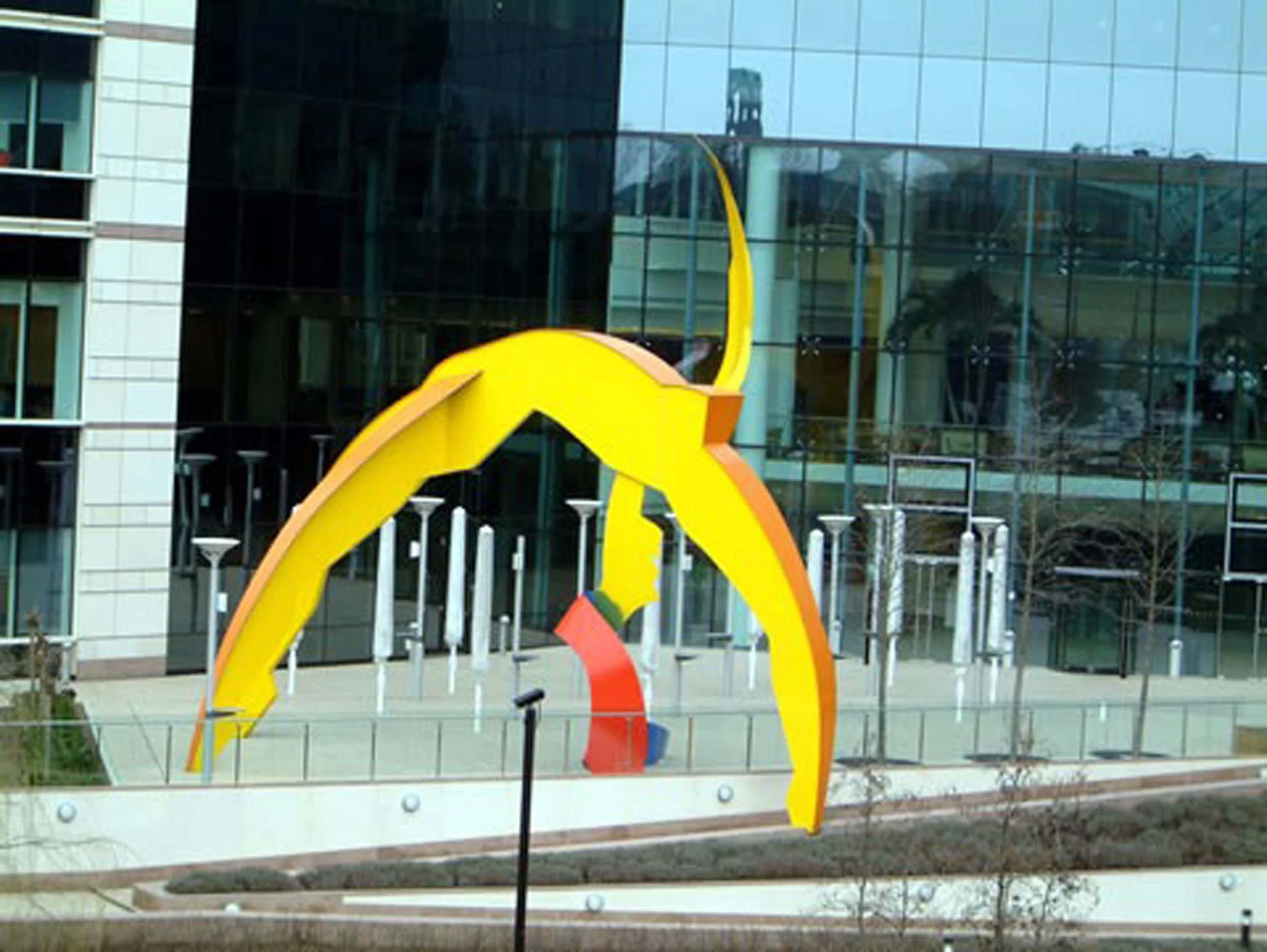 Colorful abstract sculpture of a dinosaur, situated outside a modern glass building with reflections of trees and sky.