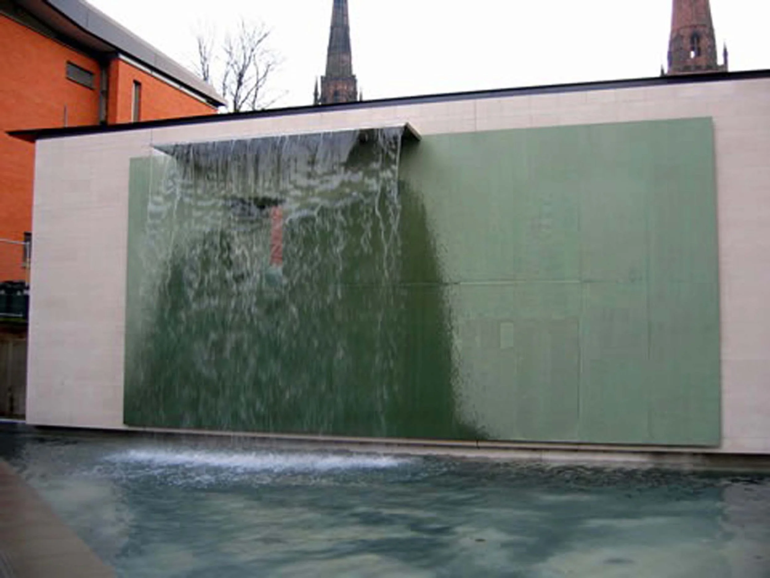 A modern outdoor water feature with water cascading from a tipped metallic spout onto a textured green wall, with trees and church steeples visible in the background.