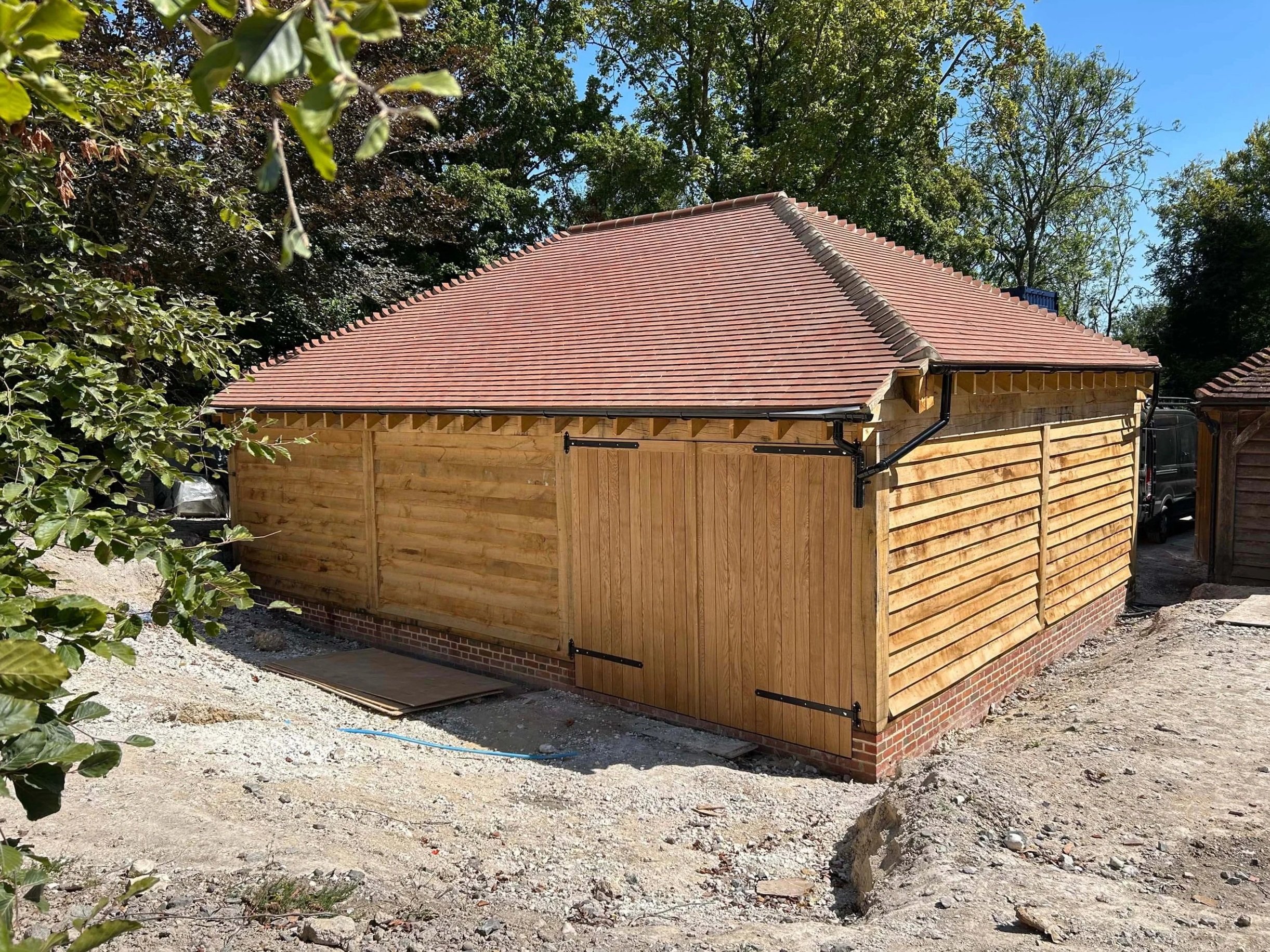 A newly constructed wooden garage with a red tiled roof, surrounded by trees and dirt ground.