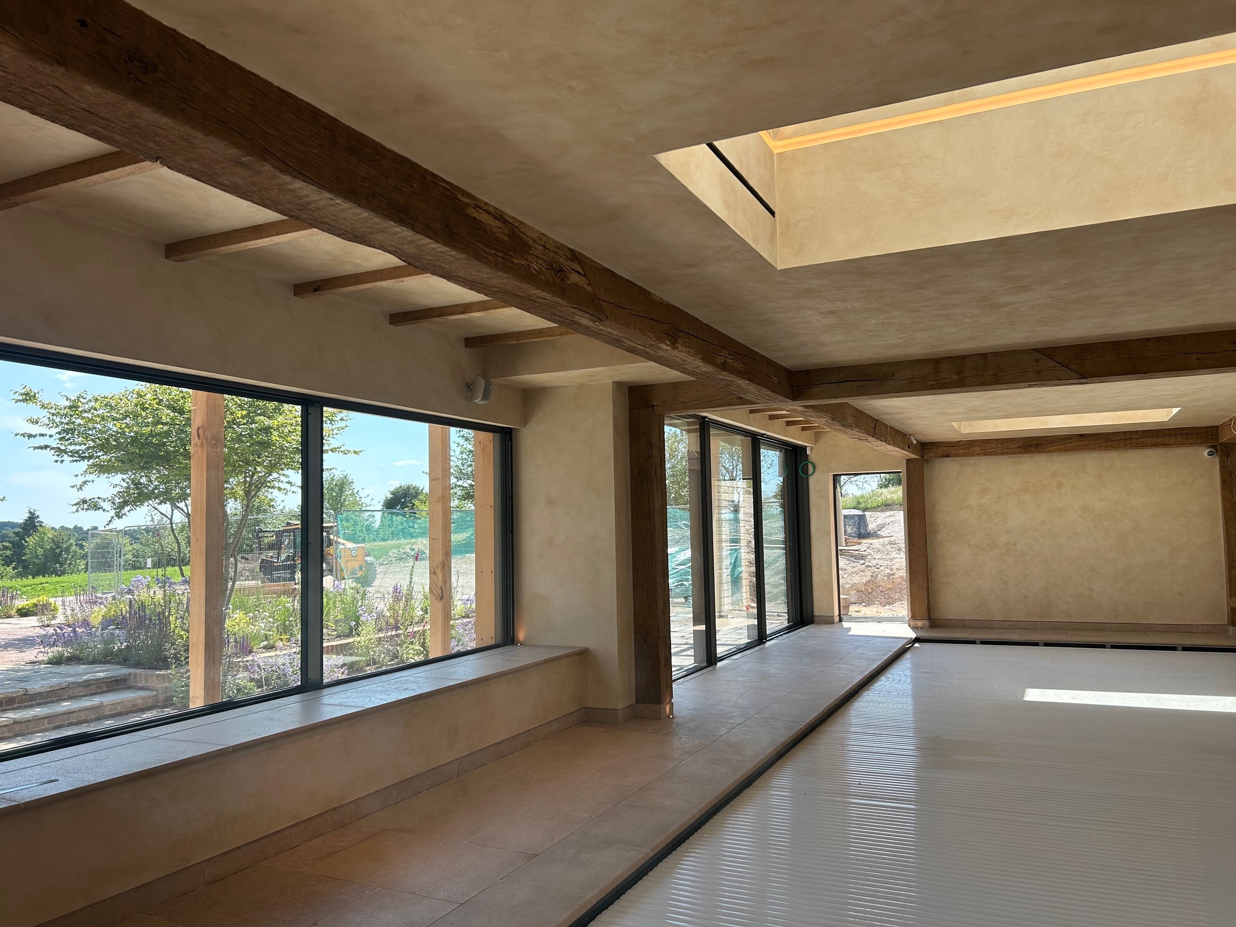 Empty interior room with large glass windows, wooden beams, and a view of a garden outside.