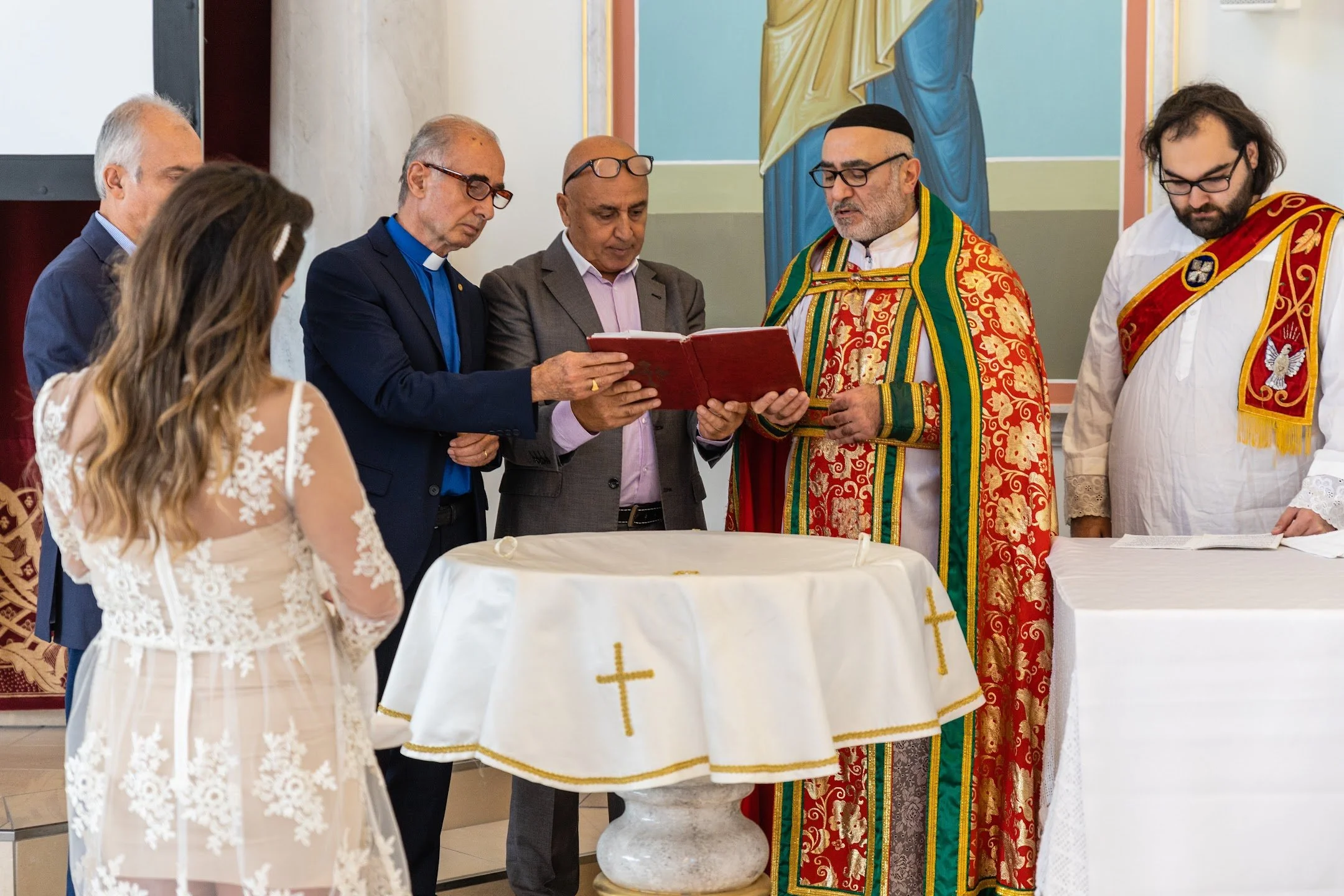 A wedding ceremony with a couple standing before a priest and assisting clergy. The bride is in a white lace dress, and the groom is in a grey suit. The clergy members are dressed in colorful liturgical vestments, and all individuals are gathered around an altar covered with a white cloth with gold crosses.