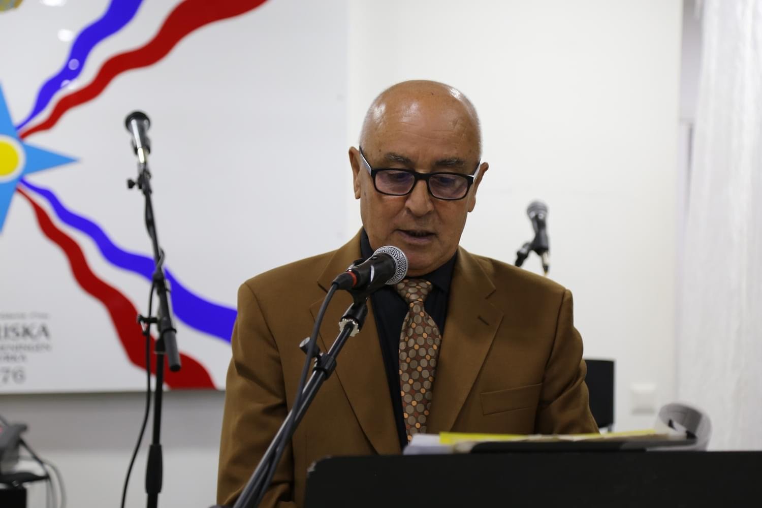 A man with glasses, dressed in a brown suit and tie, is speaking into a microphone at a podium during a speech or presentation.