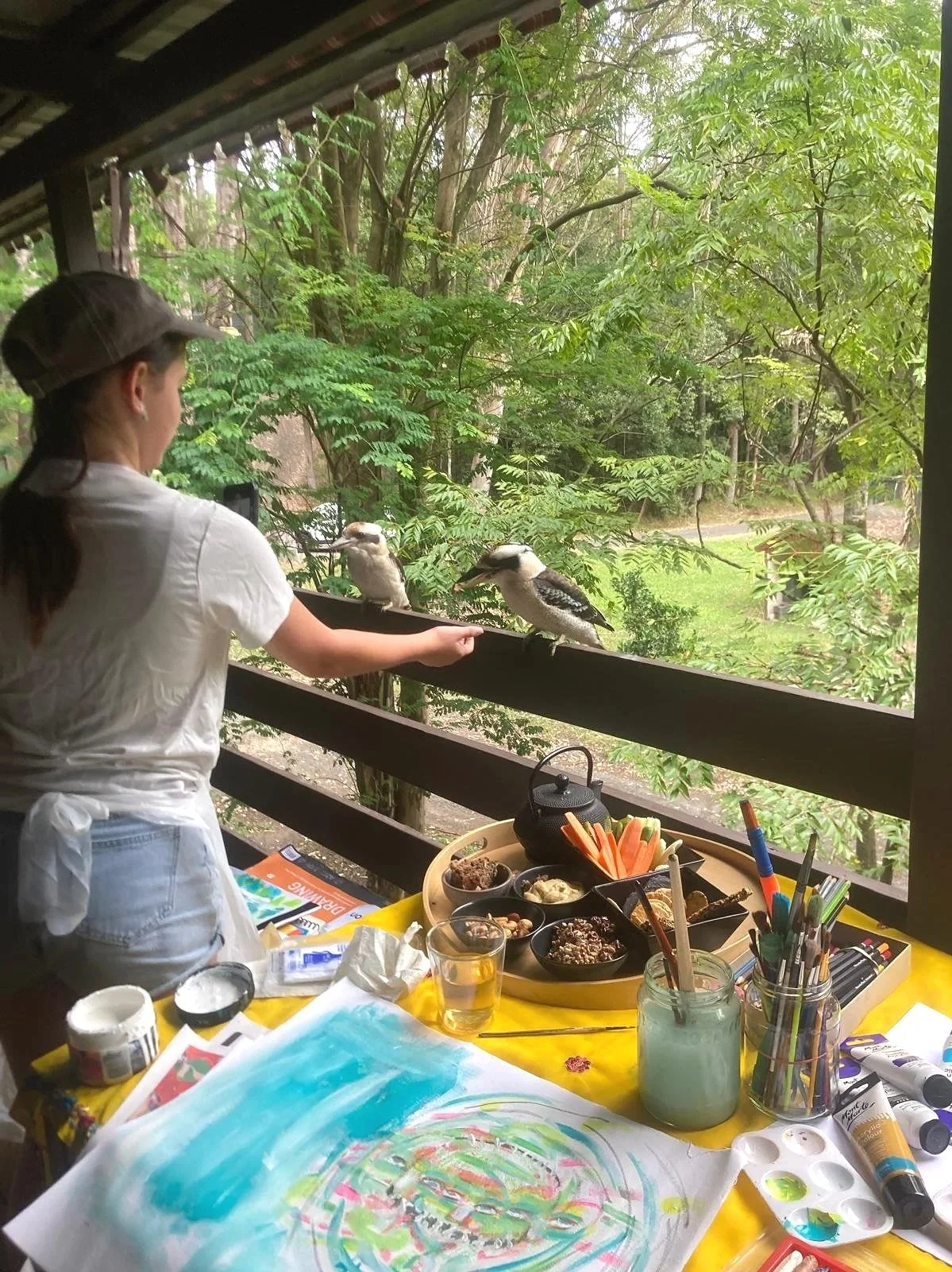 A person wearing a white shirt, white apron, and a brown cap standing on a covered porch, feeding two kookaburra birds perched on a black railing. The table in front has art supplies, a glass of water, jars, and a colorful abstract painting.