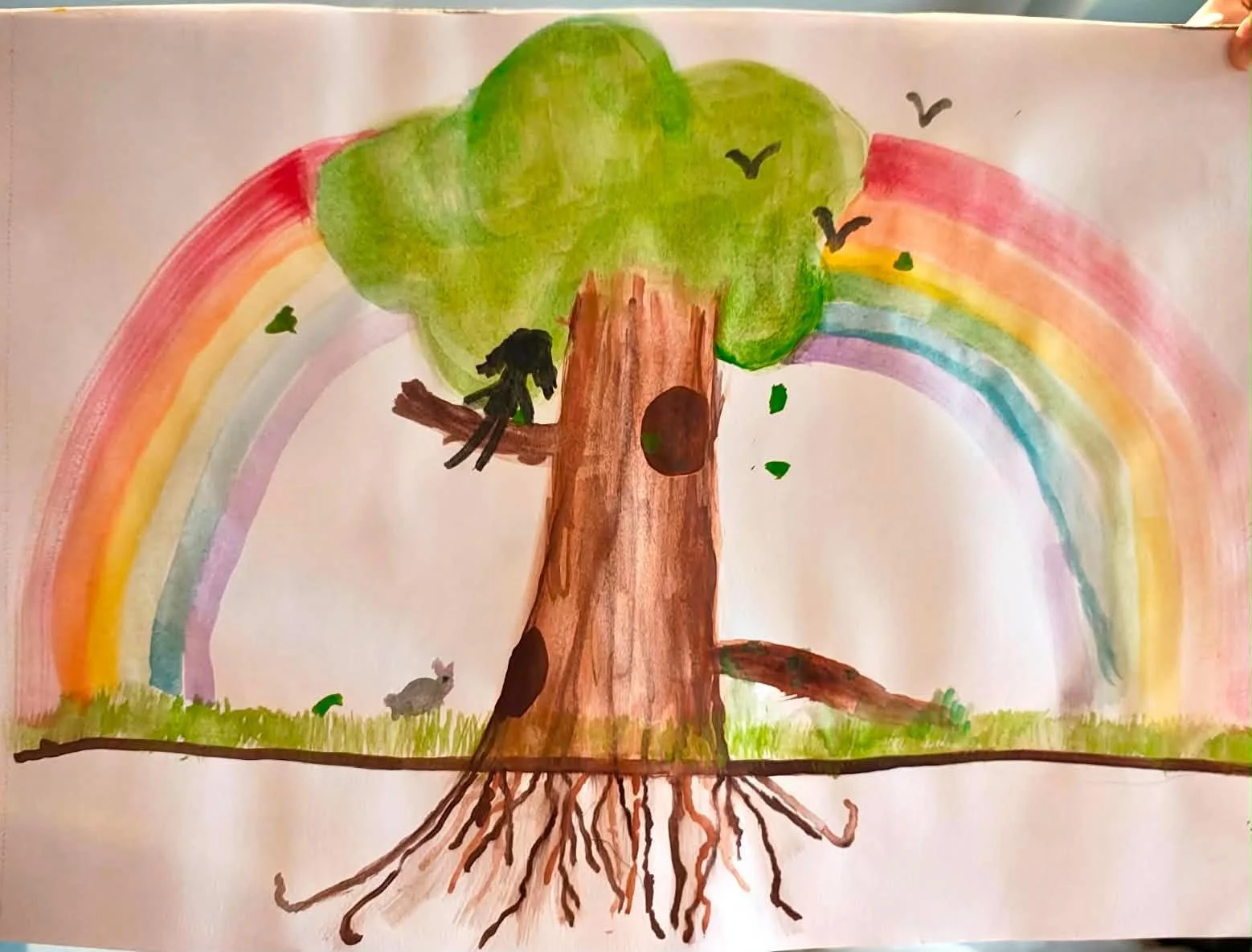 Child's drawing of a large tree with a rainbow in the background, small animals on the ground, and birds flying.