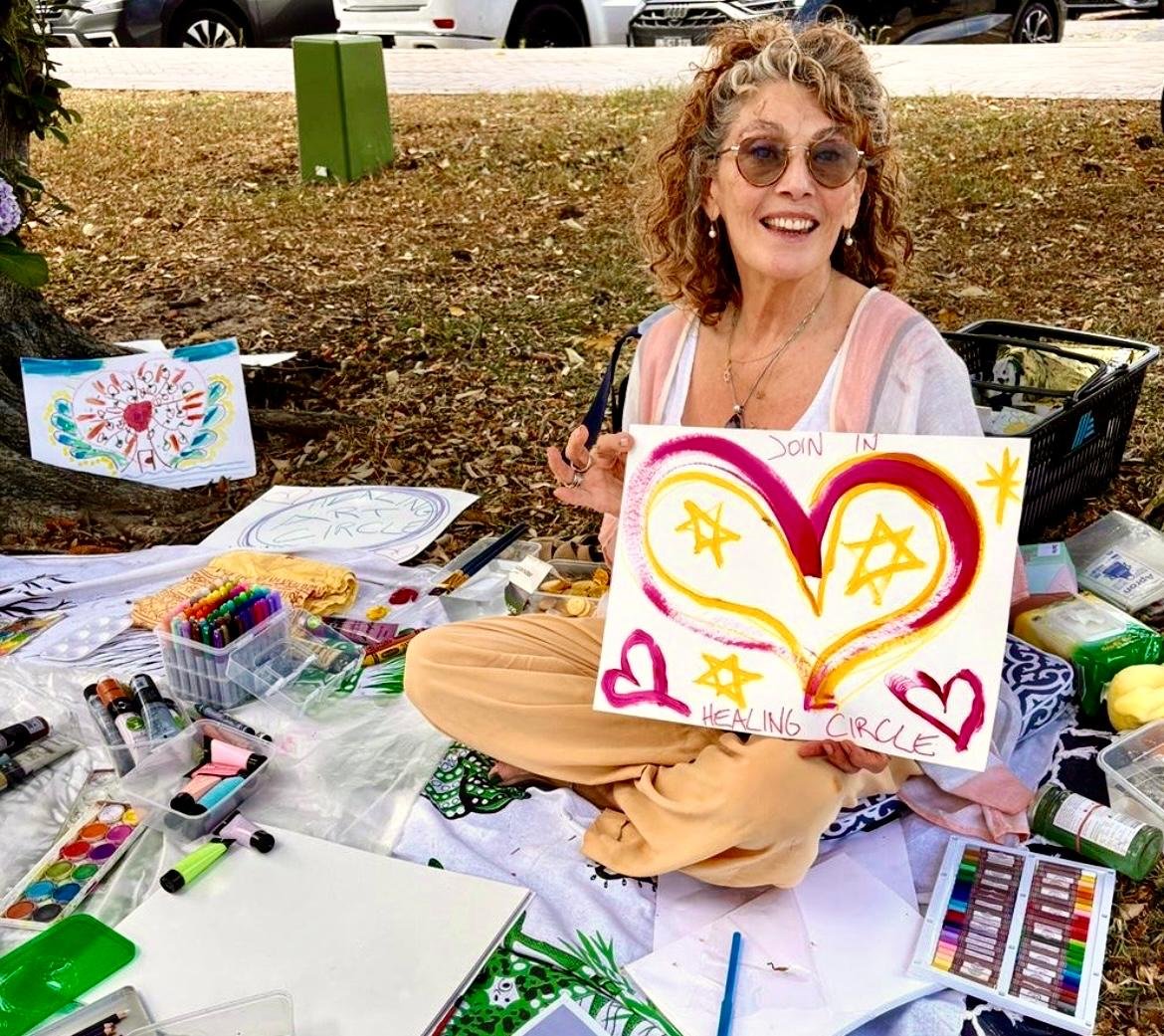 A woman sitting on the ground at an outdoor art table, holding a colorful painted sign that reads "Healing Circle" with a heart and star design. She's wearing glasses, earrings, and a light-colored top. Art supplies including markers, paints, and pap