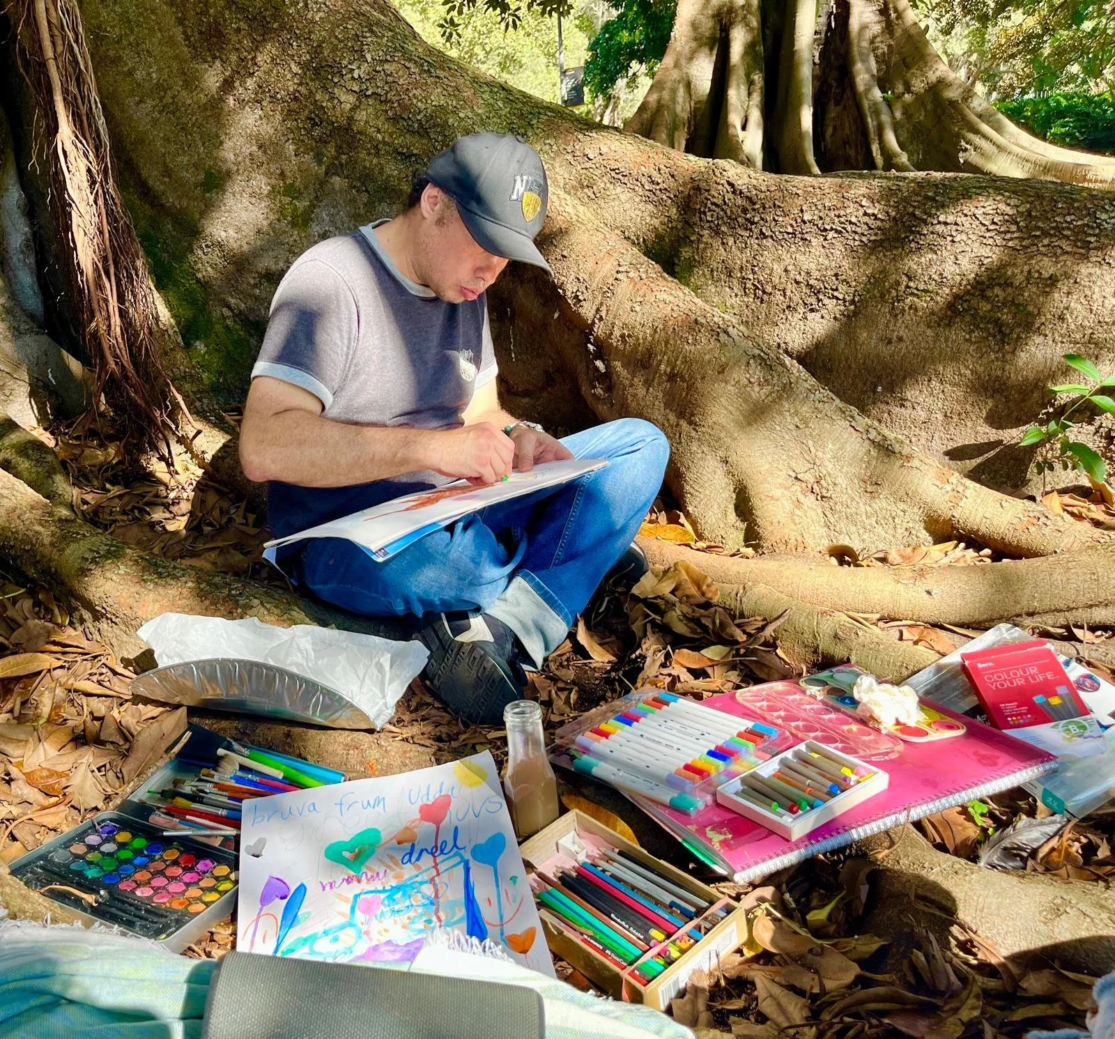 A young man sitting on the ground outdoors, drawing with markers, surrounded by art supplies such as colored pens, paint sets, and paper, beneath a large tree with sprawling roots.
