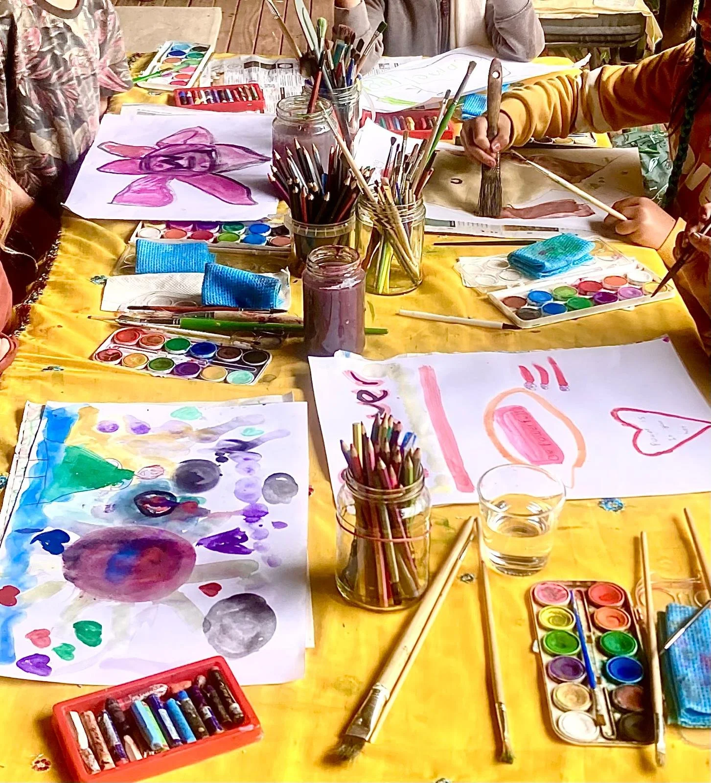 Children's watercolor painting activity with various paints, brushes, and paper on a yellow table.