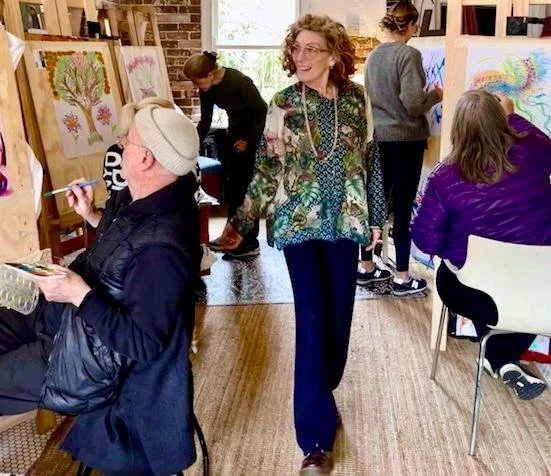 A group of people participating in an art class, with some drawing on canvases and others observing or walking around, in a cozy indoor setting with natural light and colorful artwork displayed.