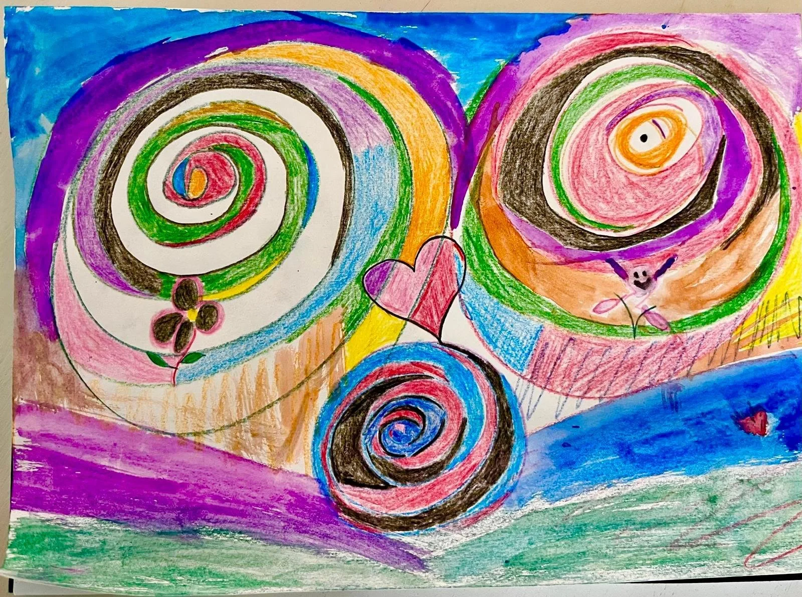 Colorful abstract drawing with swirls, hearts, flowers, and a smiling face, created with crayons or colored pencils.