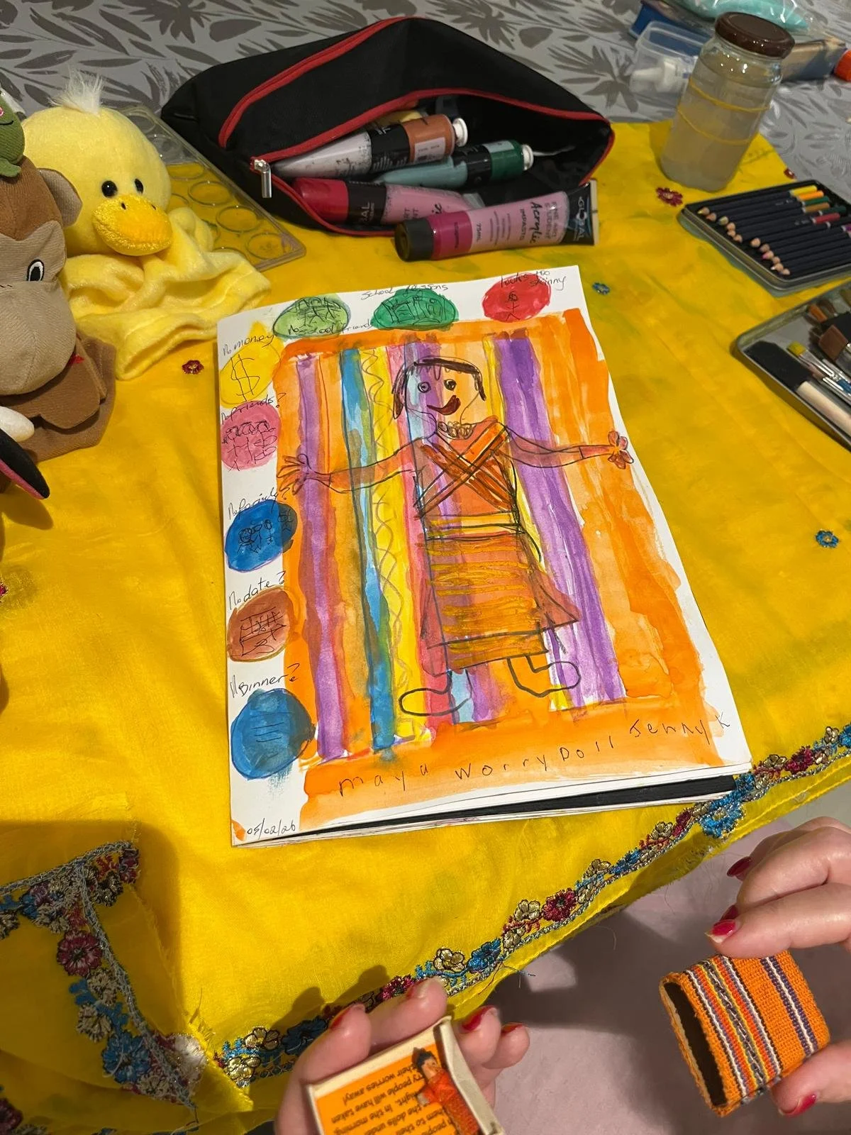 A colorful child's drawing of a woman with her arms outstretched, surrounded by a rainbow background and various colored circles labeled with words. The drawing is on a piece of paper on a yellow cloth table, with art supplies nearby, including paint