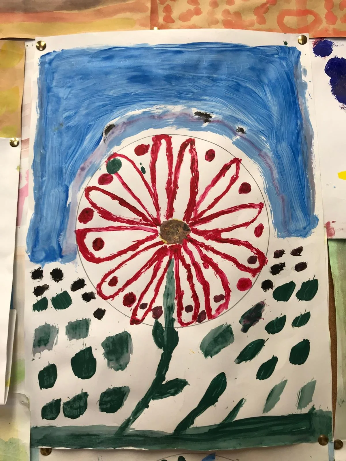 Children's artwork depicting a large flower with red petals and a brown center, green stem and leaves, set against a background of blue sky and scattered green and black dots.