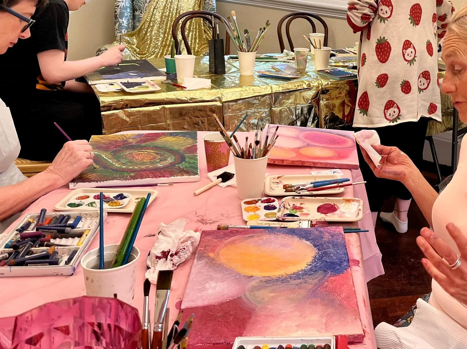 People painting on canvases at a social art event, with paint supplies and brushes on the table.