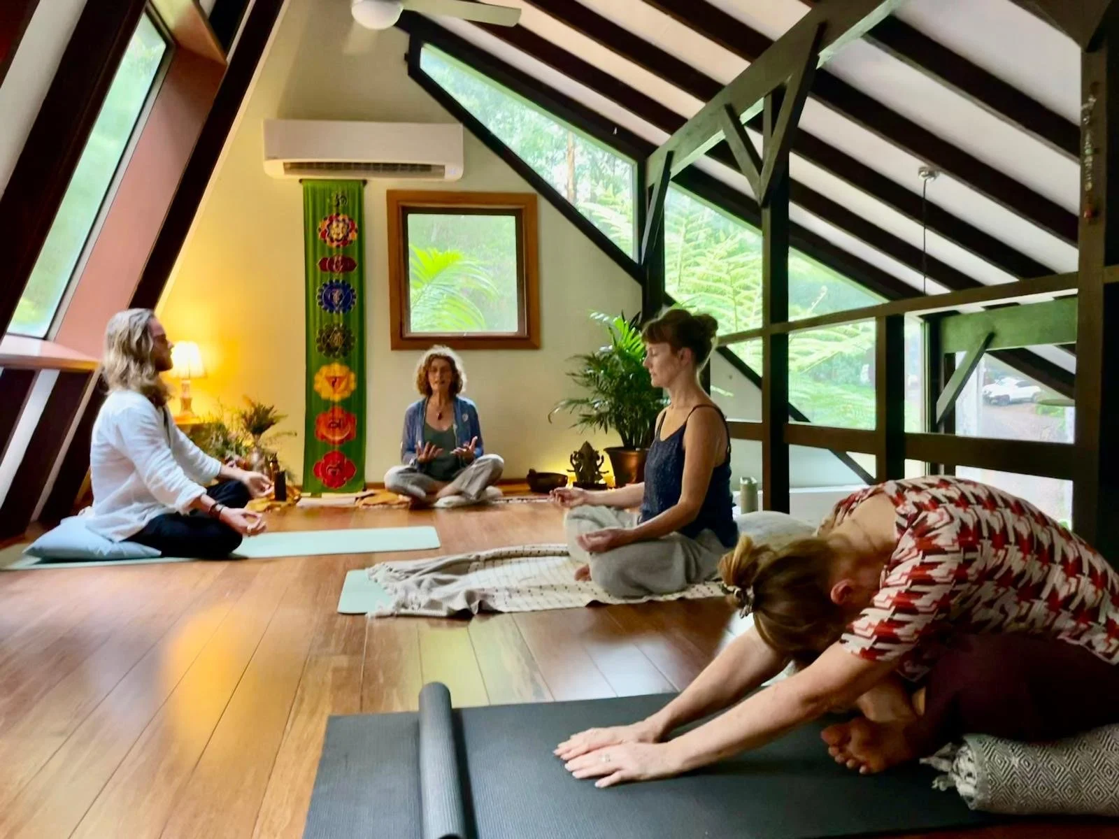 Group of five people participating in a yoga or meditation session in a spacious, well-lit room with wooden floors, angled windows, and greenery visible outside. They are seated or lying on mats, engaging in poses or relaxation. One person appears to