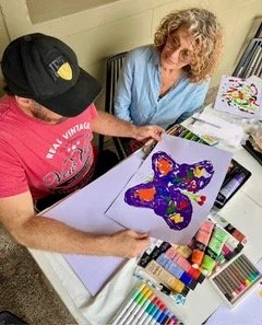 Two women sitting at a table with art supplies, working on a painted butterfly canvas.
