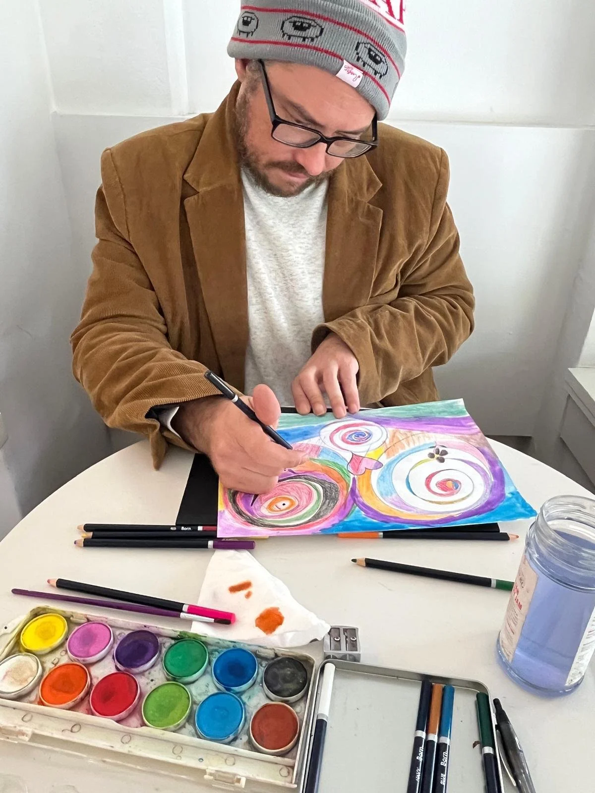 A man is sitting at a white table and painting a colorful, abstract artwork with swirls and spirals using watercolors. On the table are various art supplies, including watercolor paints, brushes, and markers.