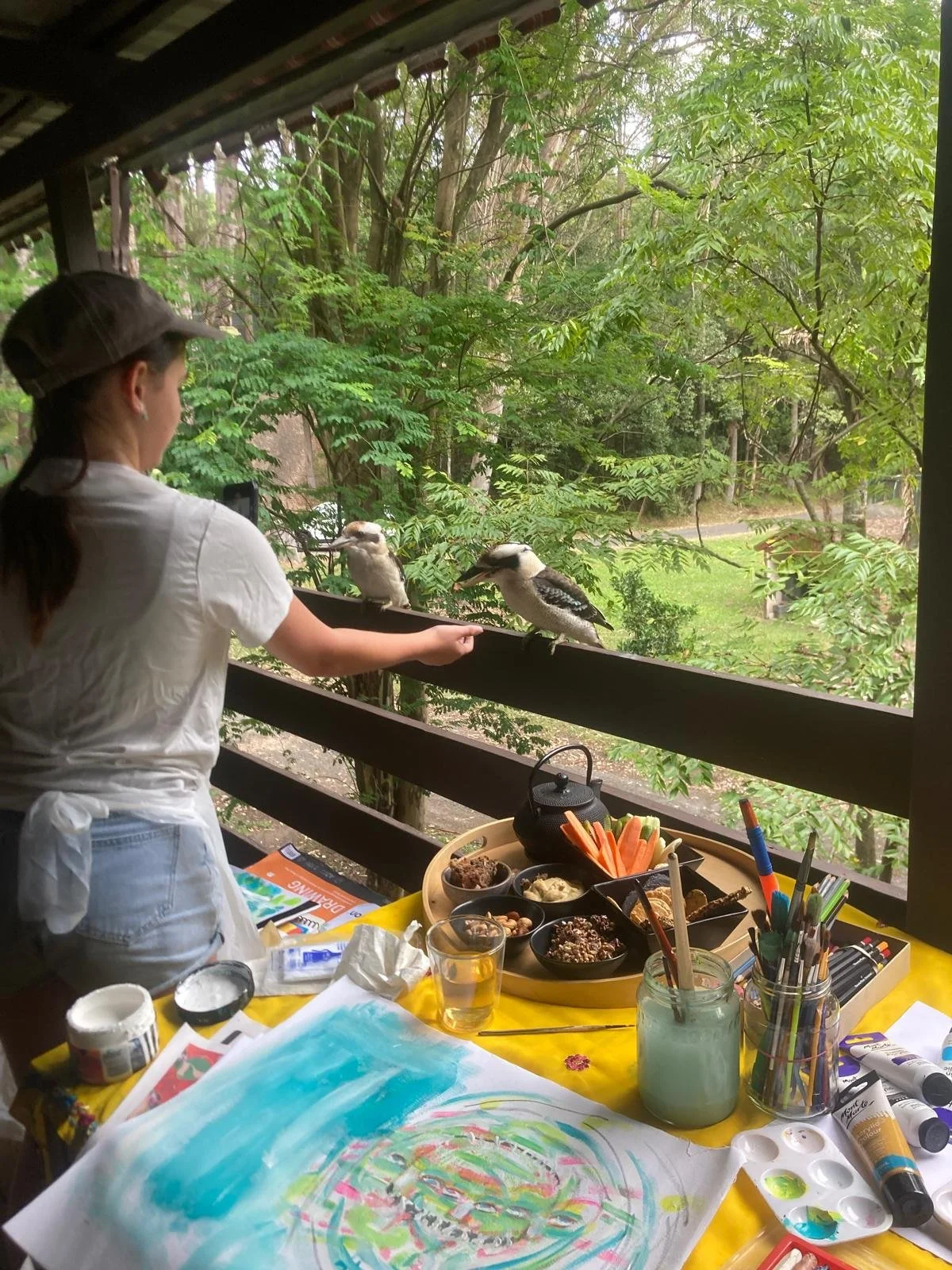 A person wearing a white shirt, apron, and cap, is on a balcony with tables of art supplies and food, feeding two kookaburra birds perched on a railing, overlooking green trees and a grassy area outside.