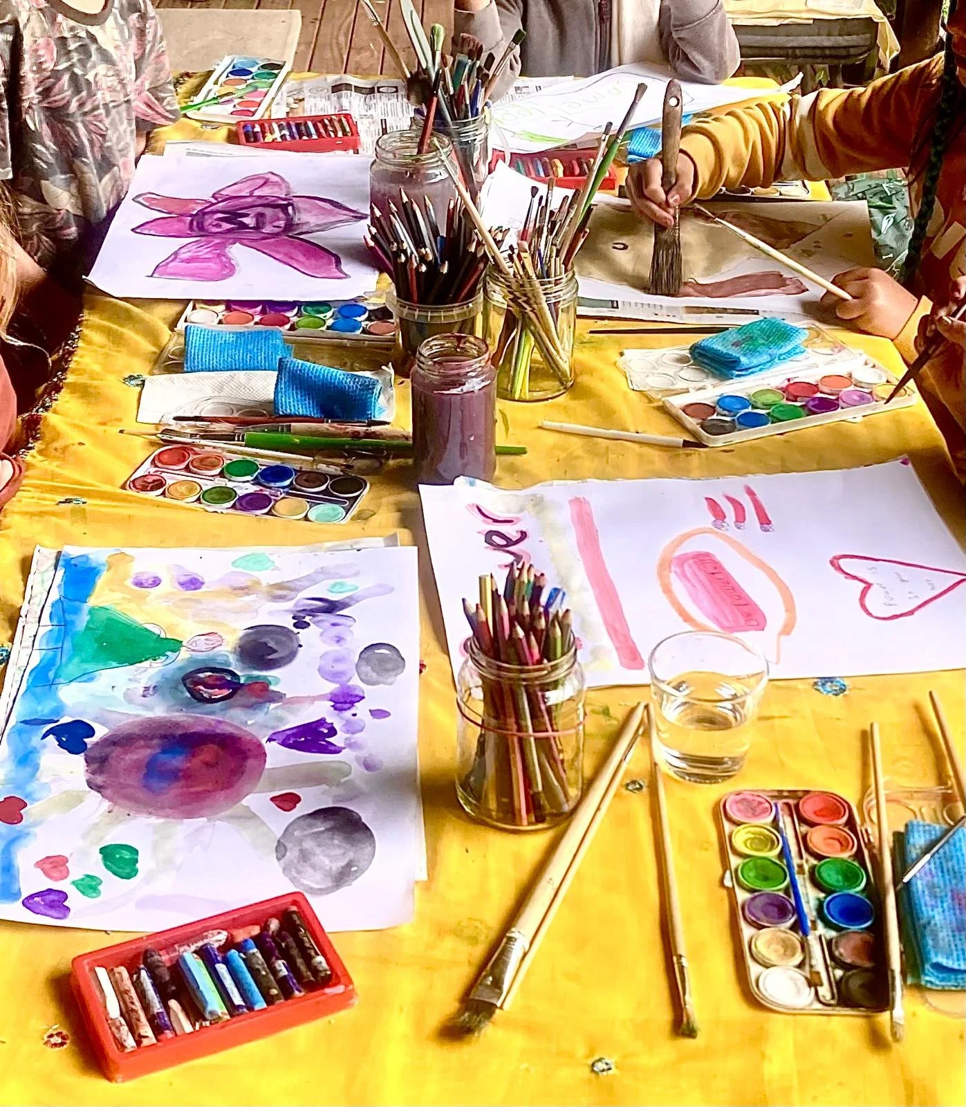 Children drawing and painting with watercolors on paper, with art supplies such as colored pencils, paintbrushes, watercolors, and water jars on a yellow table.