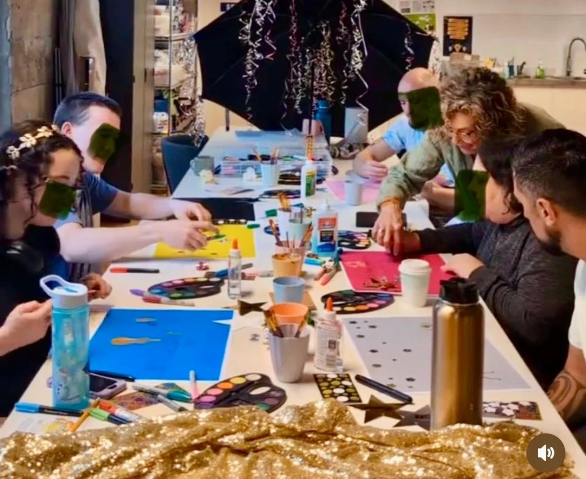 A group of people participating in a craft activity at a table with art supplies, including paints, brushes, and paper, decorated with a large black umbrella and festive gold cloth.