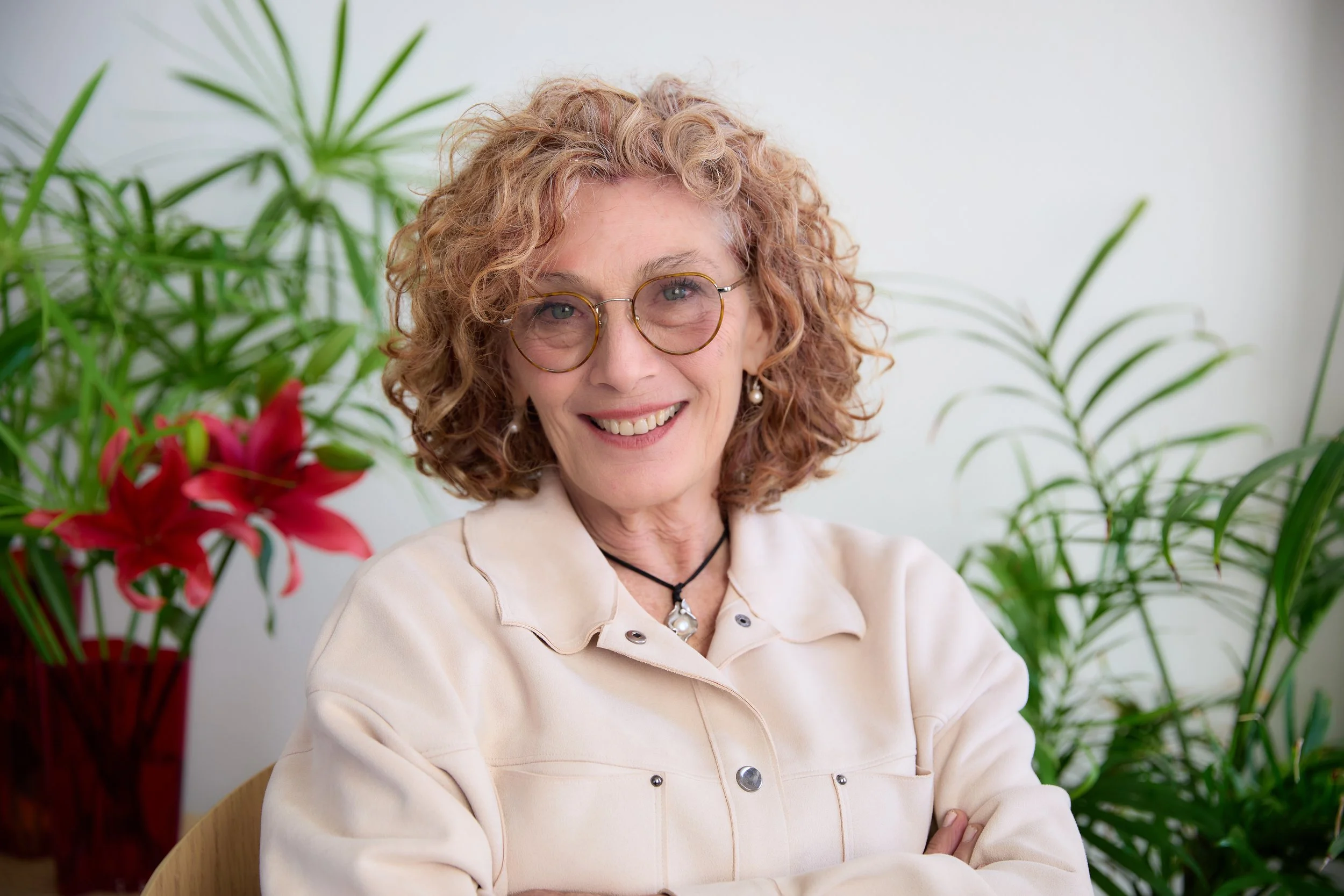 A smiling woman with curly red hair and glasses sitting near green plants with pink flowers in the background.