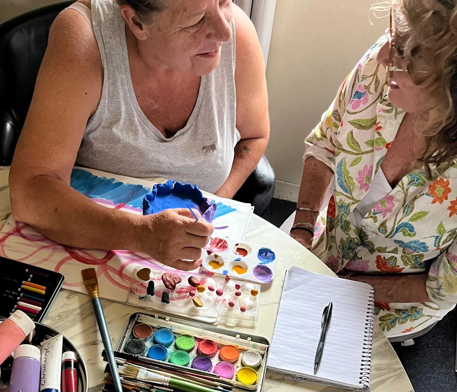 Two women are sitting at a table engaged in a painting activity with watercolors, brushes, and notebooks. The woman on the left is painting a small object, while the woman on the right observes. The table has various paints, brushes, and art supplies