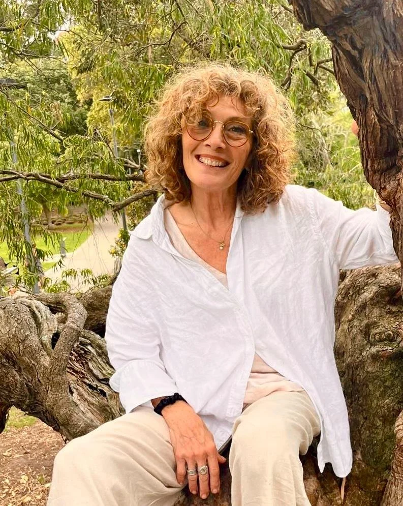 A woman with curly blonde hair and glasses sitting in a tree, smiling at the camera, surrounded by green leaves.