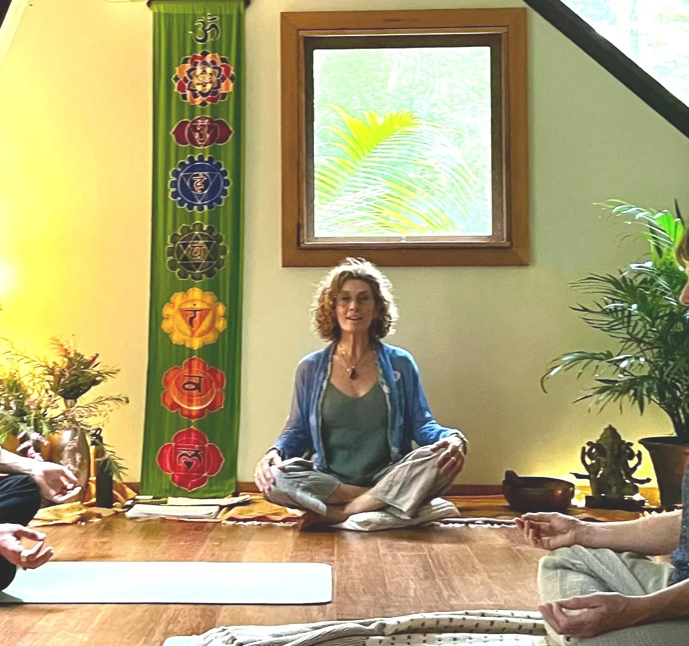 Jenni as a woman with curly hair sitting cross-legged on the floor in a room decorated with plants and a vertical banner with chakra symbols, during a yoga or meditation session.