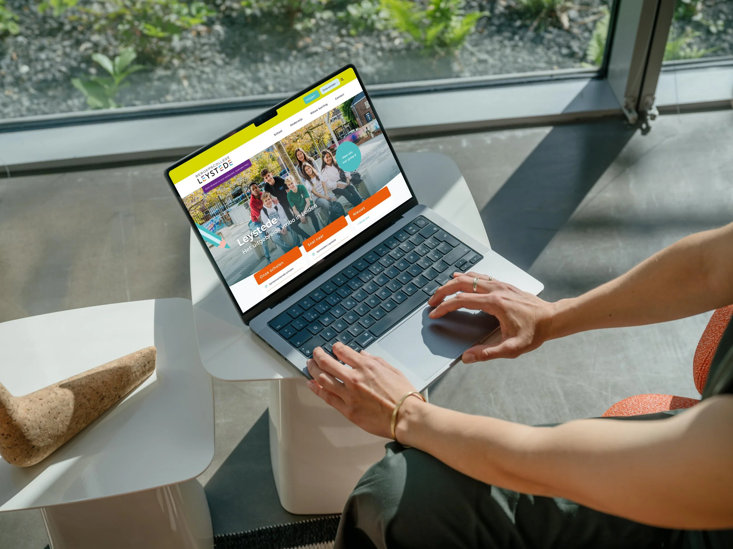 A person using a silver laptop on a white table. The person's hands are on the keyboard, wearing a gold bracelet. The laptop screen displays a colorful website