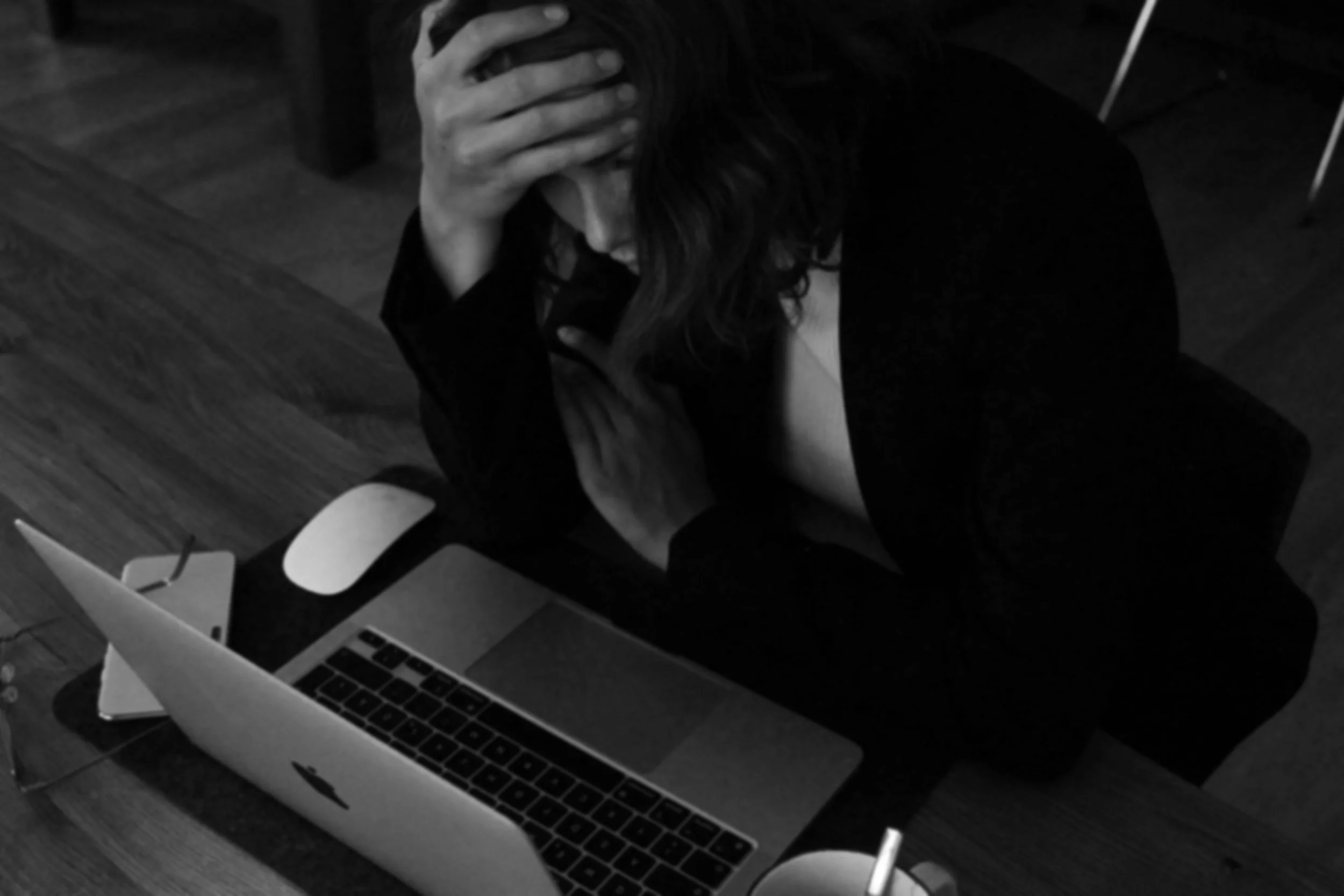 A person with long hair, wearing dark clothing, sitting at a desk with a laptop and a smartphone, holding their head with one hand in a distressed or overwhelmed manner.