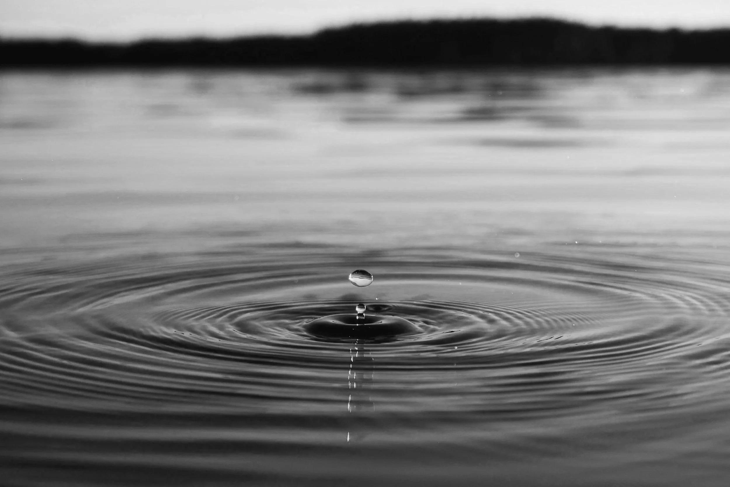 Close-up black and white photograph of a water droplet falling into a calm body of water, creating ripples representing what trauma can do.