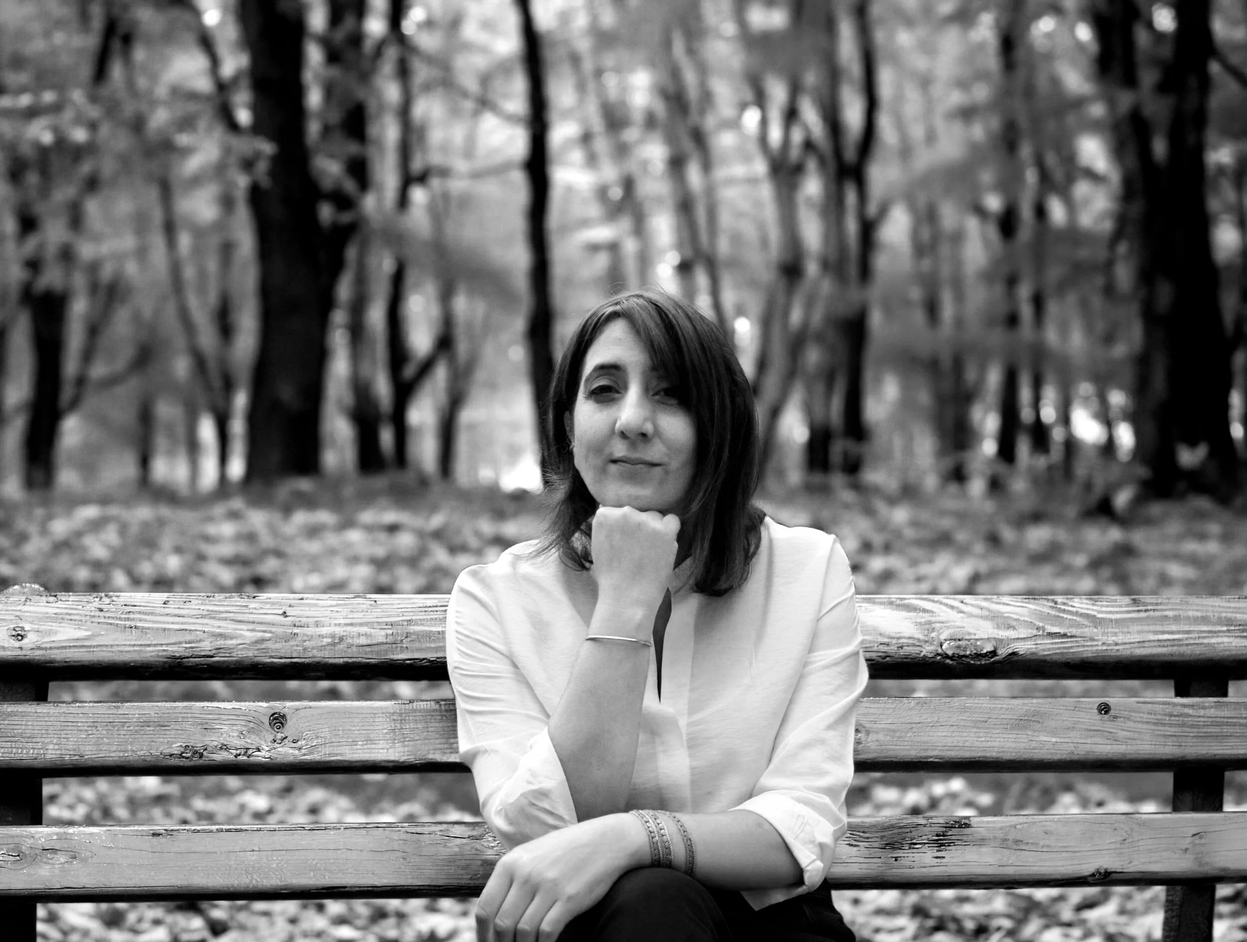 A black and white photo of a woman sitting on a wooden park bench made of horizontal planks, outdoors with trees in the background. The woman has shoulder-length hair and is wearing a light-colored long-sleeve top. She has a subtle smile and is resting her chin on her fist, looking at the camera.