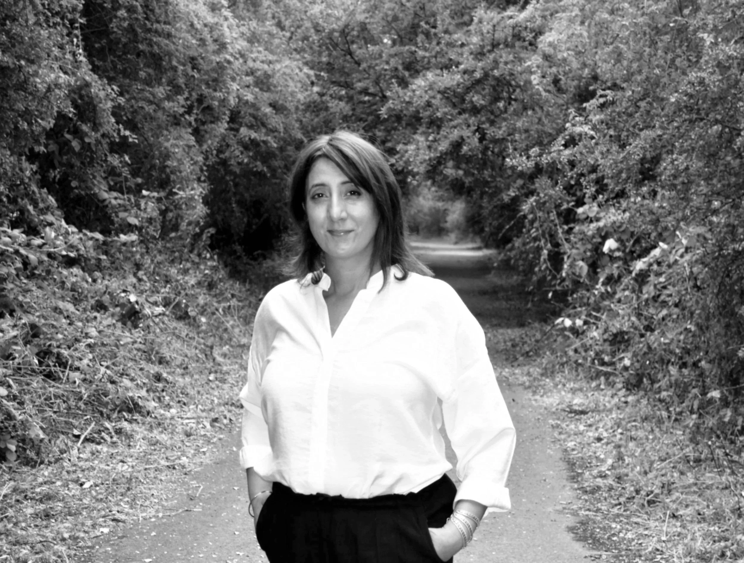 A woman with shoulder-length hair, wearing a white shirt, stands on a forest path with trees on both sides.