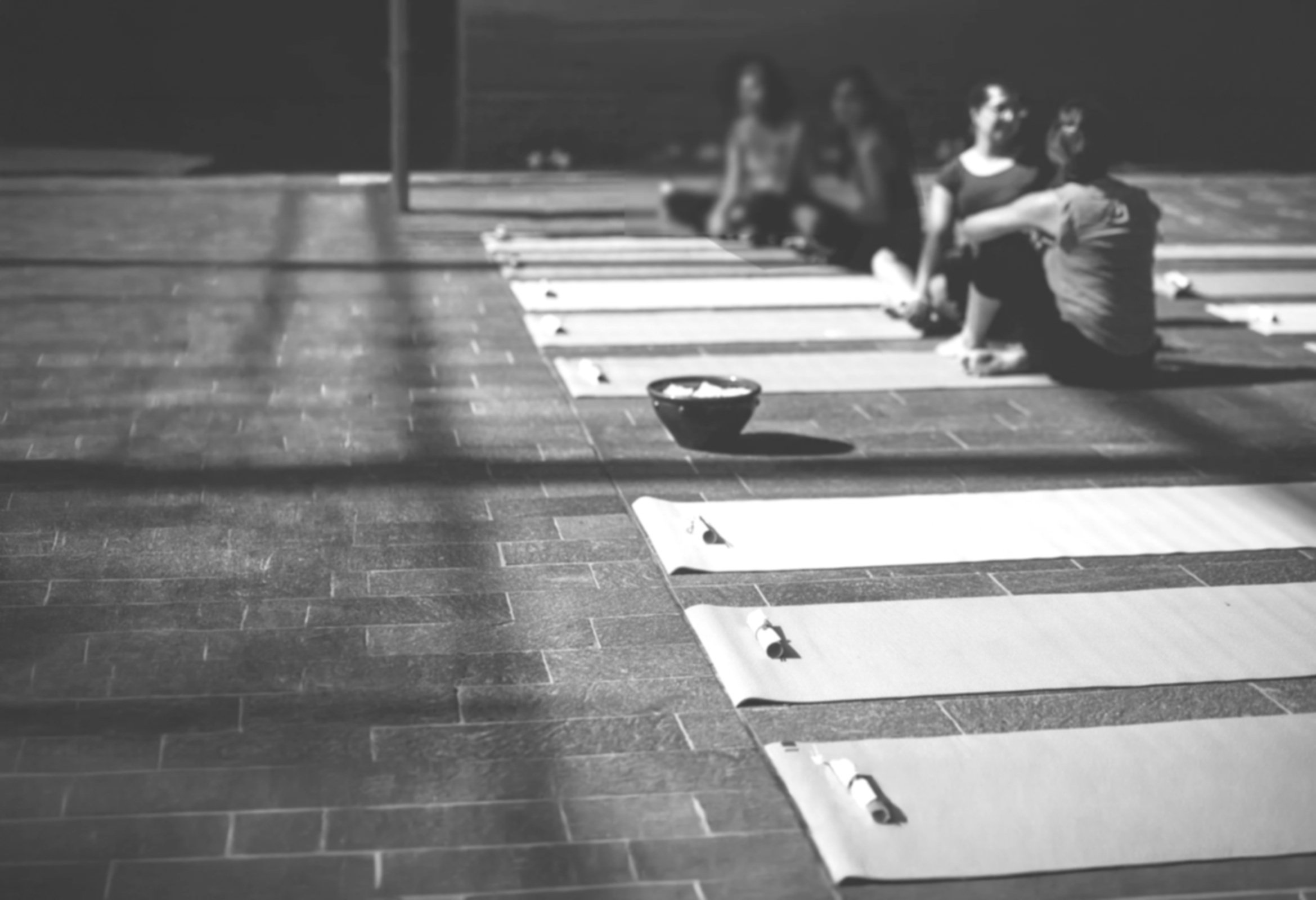 People sitting cross-legged on the floor behind mats for a yoga class or meditation session, with a bowl in the foreground, in a dark indoor space.