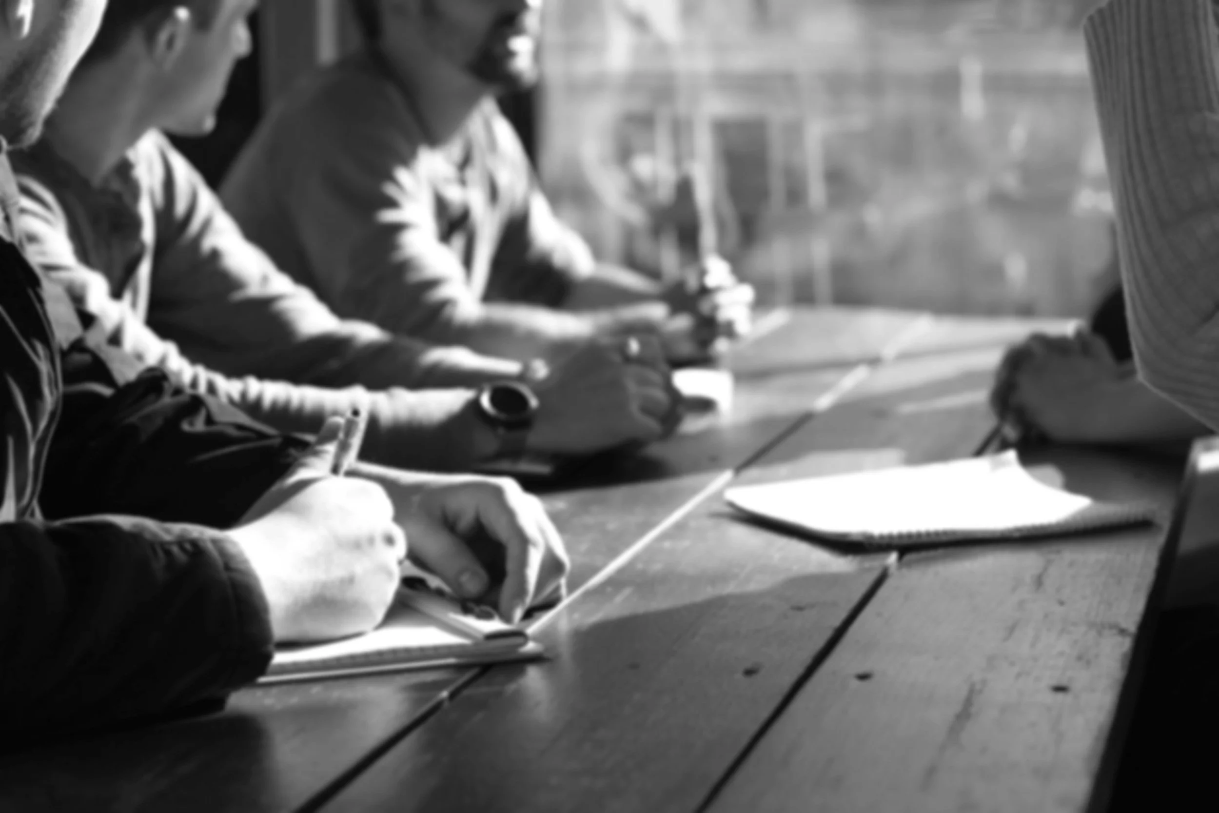 Black and white photo of three people sitting at a table, writing or drawing on paper, with notebooks and pens.