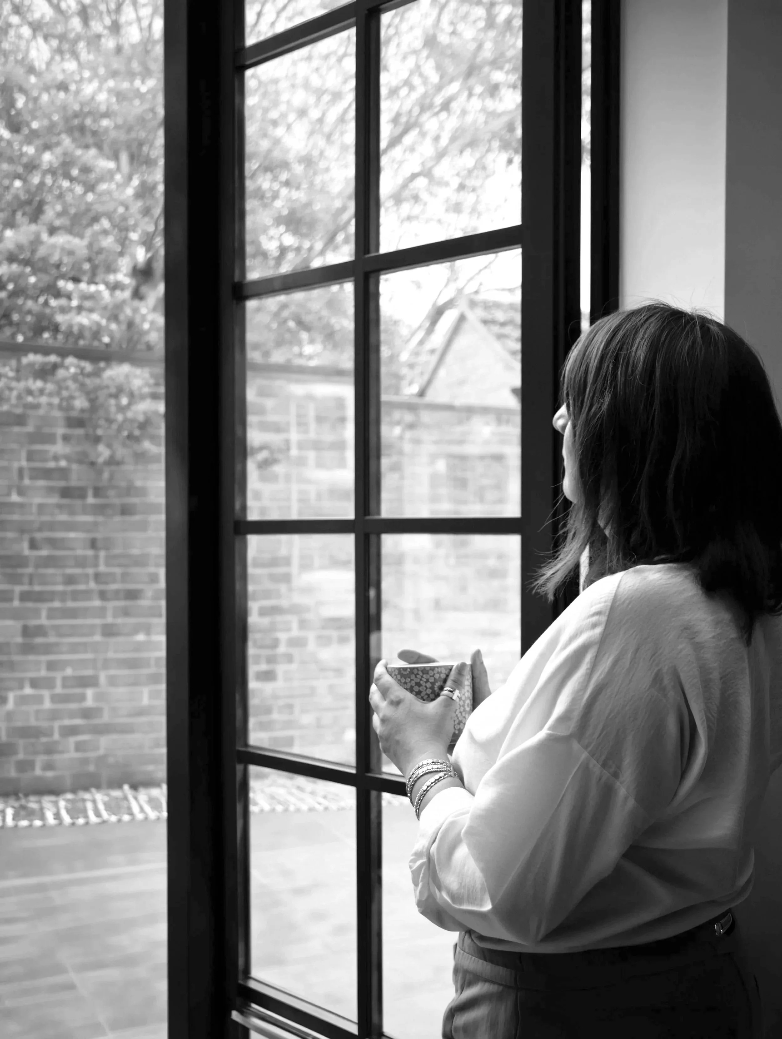 A woman with shoulder-length hair, wearing a light-colored blouse and jewelry, stands near a large window with a grid-like frame, holding a small patterned cup, with an outdoor scene including trees and a brick wall visible through the window.