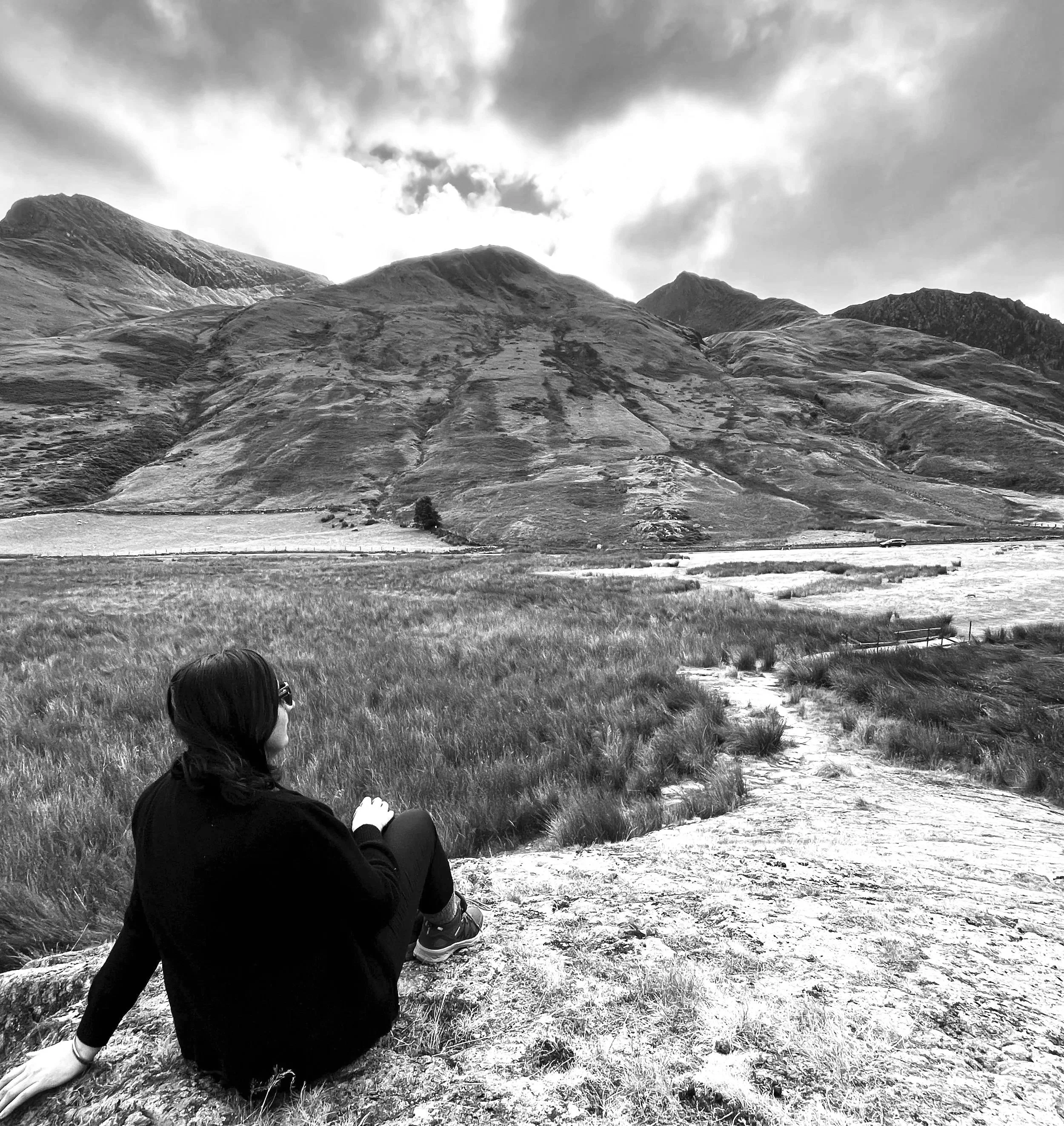 A person sitting on grass in front of a mountain landscape, wearing a black jacket, with a ponytail, looking to the left. The terrain has grass and a pathway, with mountains and a cloudy sky in the background.