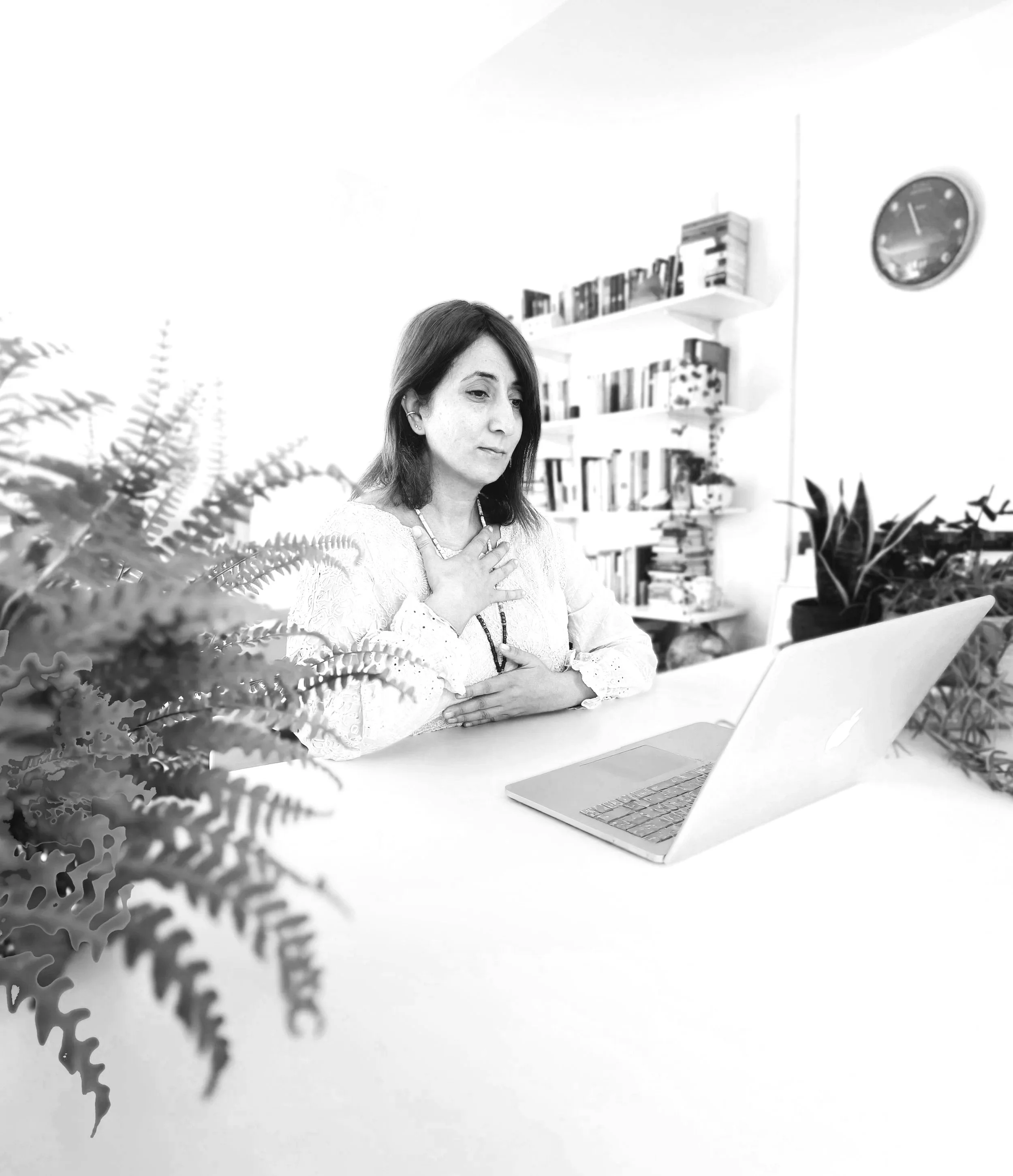 A woman with dark hair sitting at a desk with an open laptop, surrounded by plants and bookshelves in the background, in a well-lit room working somatically with a client online.