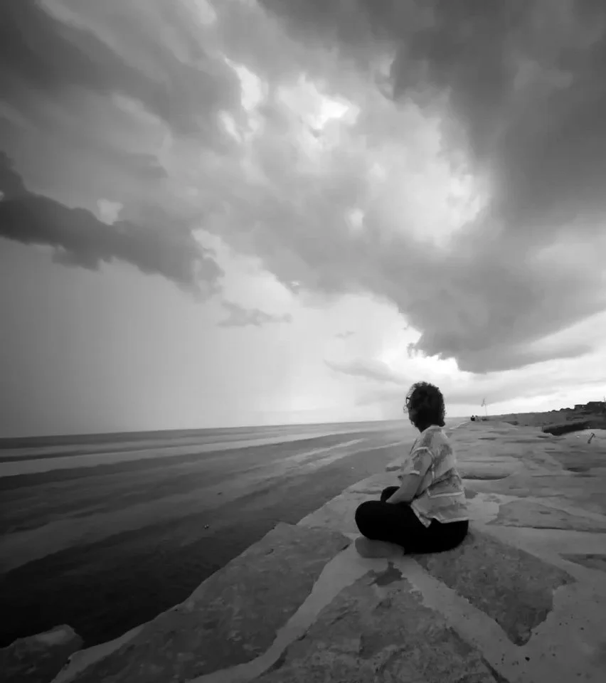 A person sitting cross-legged on a concrete pier looking at a stormy sky over the ocean in black and white representing the extent of grief.