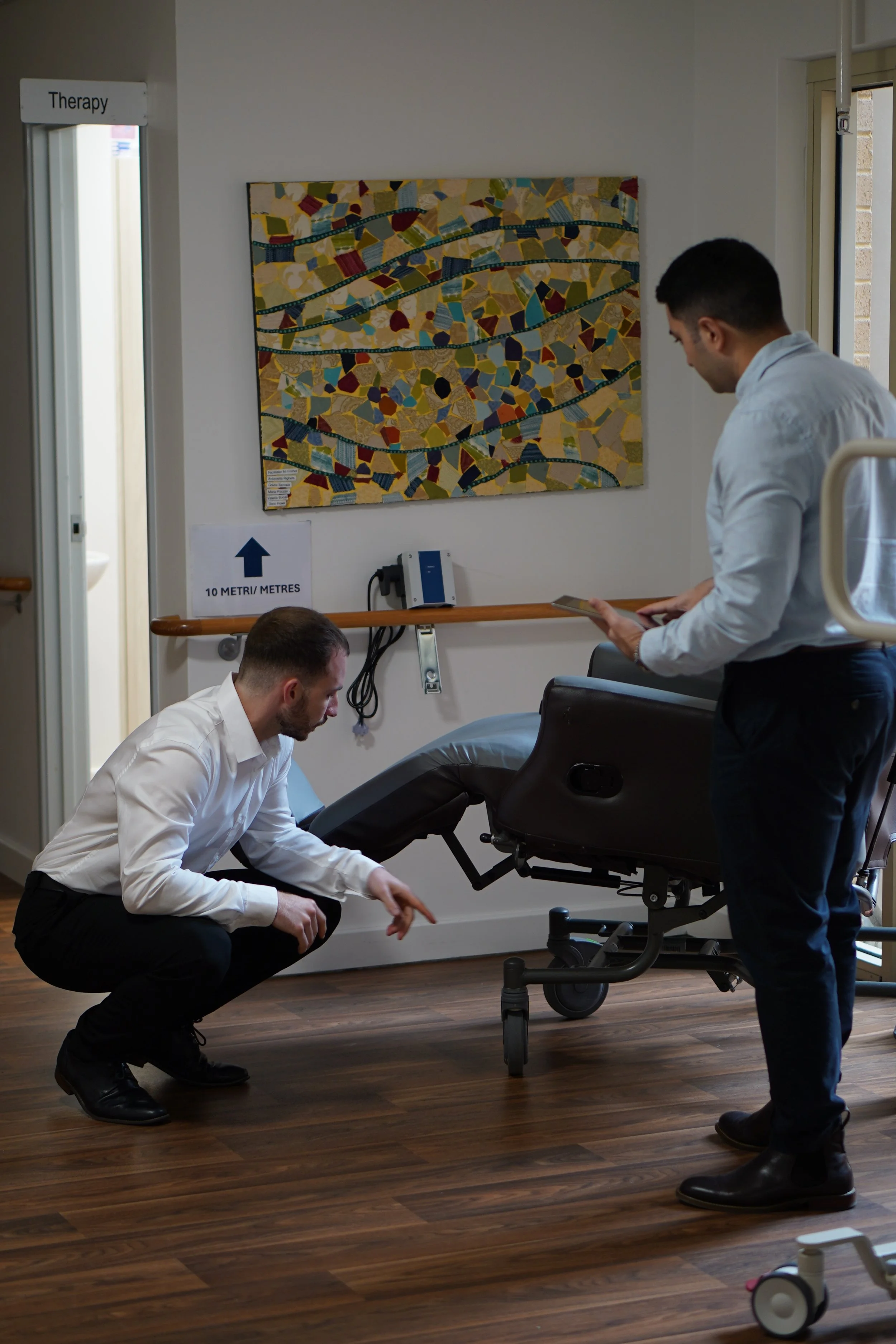 Two men in white shirts and black pants are practicing squats in a medical or therapy room. One is kneeling and touching the floor, the other is standing and holding a tablet. There is a colorful mosaic artwork on the wall and a sign indicating a 10-meter distance.