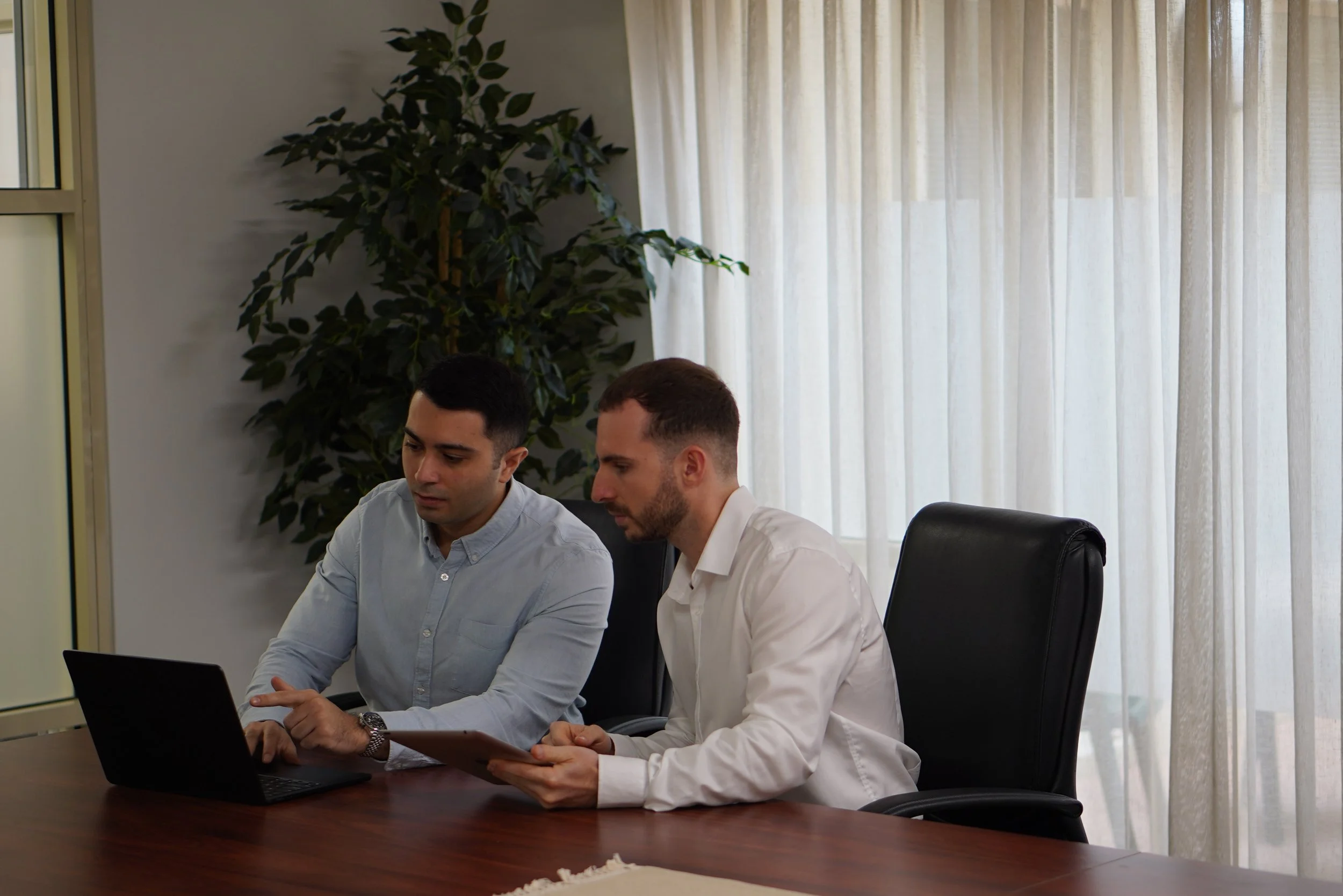 Two men sitting at a conference table in a meeting room, looking at a tablet and a laptop, with a green potted plant and beige curtains in the background.