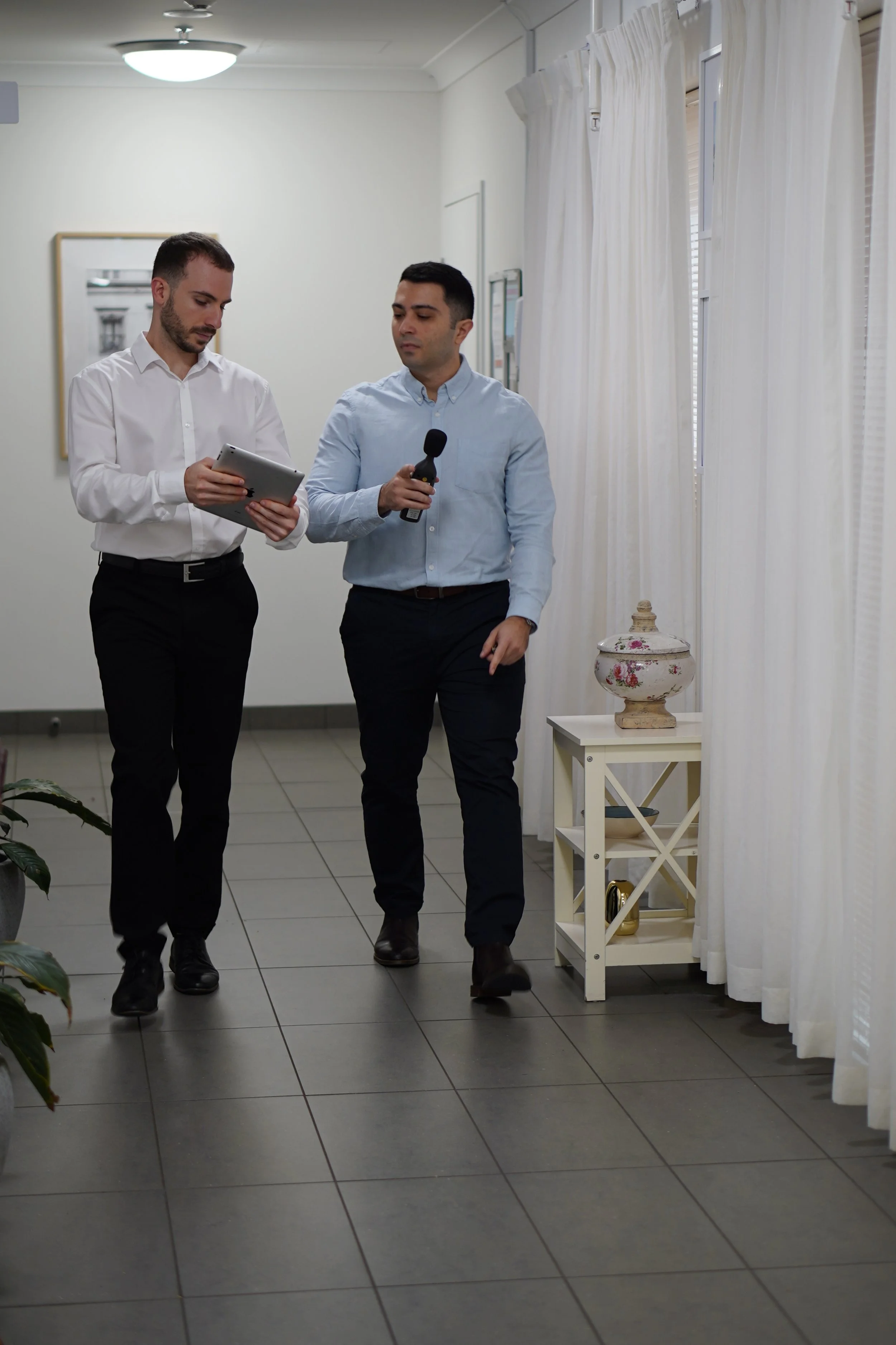 Two men in business attire walking in an office or corporate setting, one holding a microphone and the other holding a tablet, with white curtains and decorative items in the background.