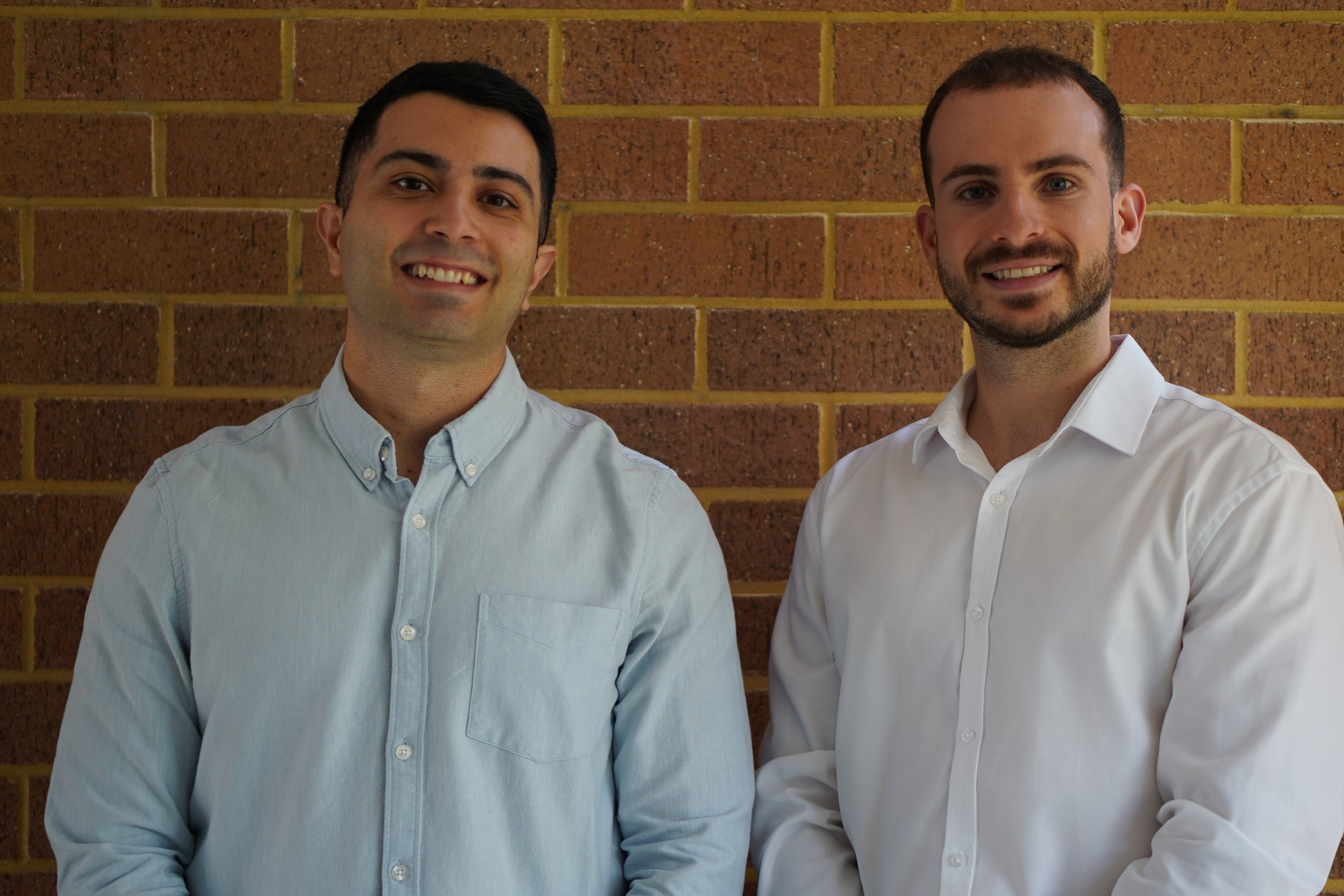 Two men smiling and standing in front of a brick wall, dressed in collared shirts.